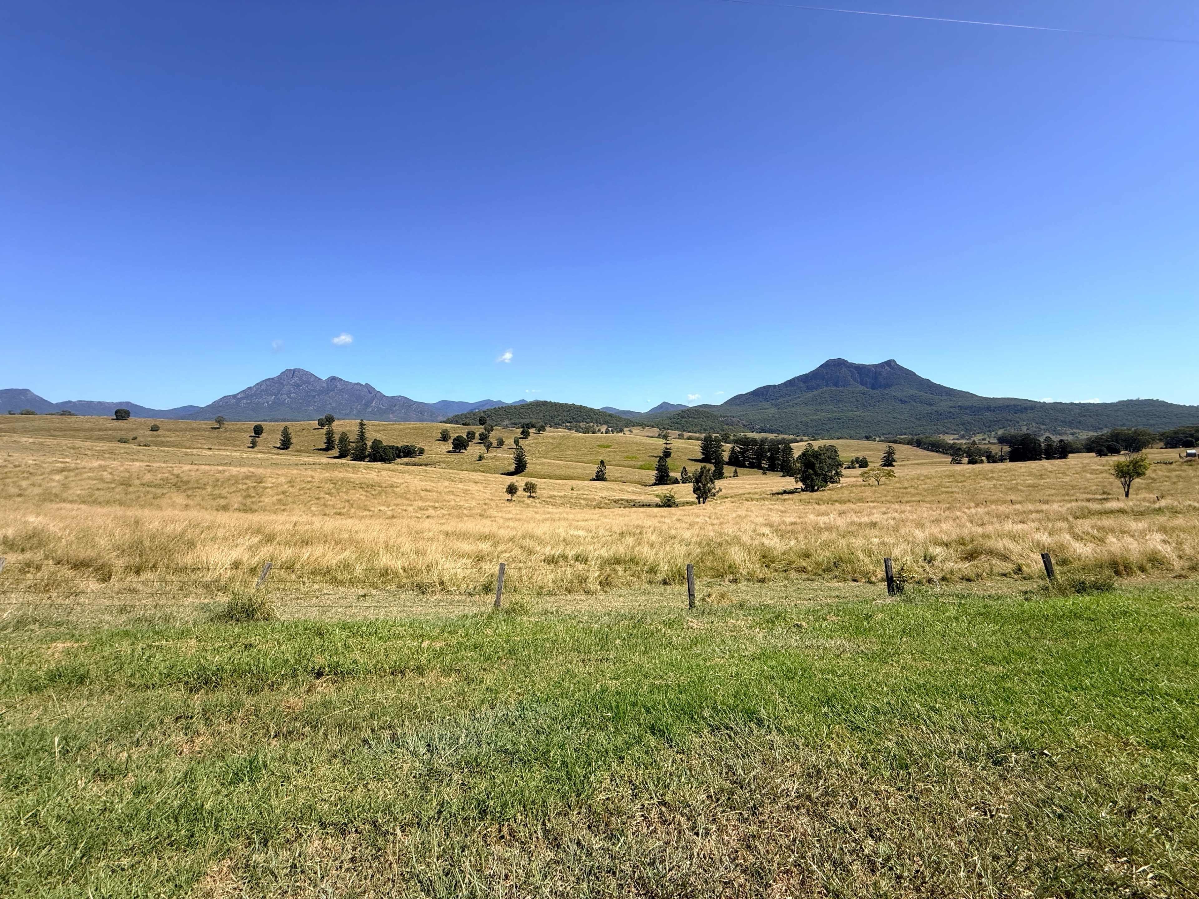 The image shows a wide landscape of grassy fields with distant mountains under a clear blue sky.