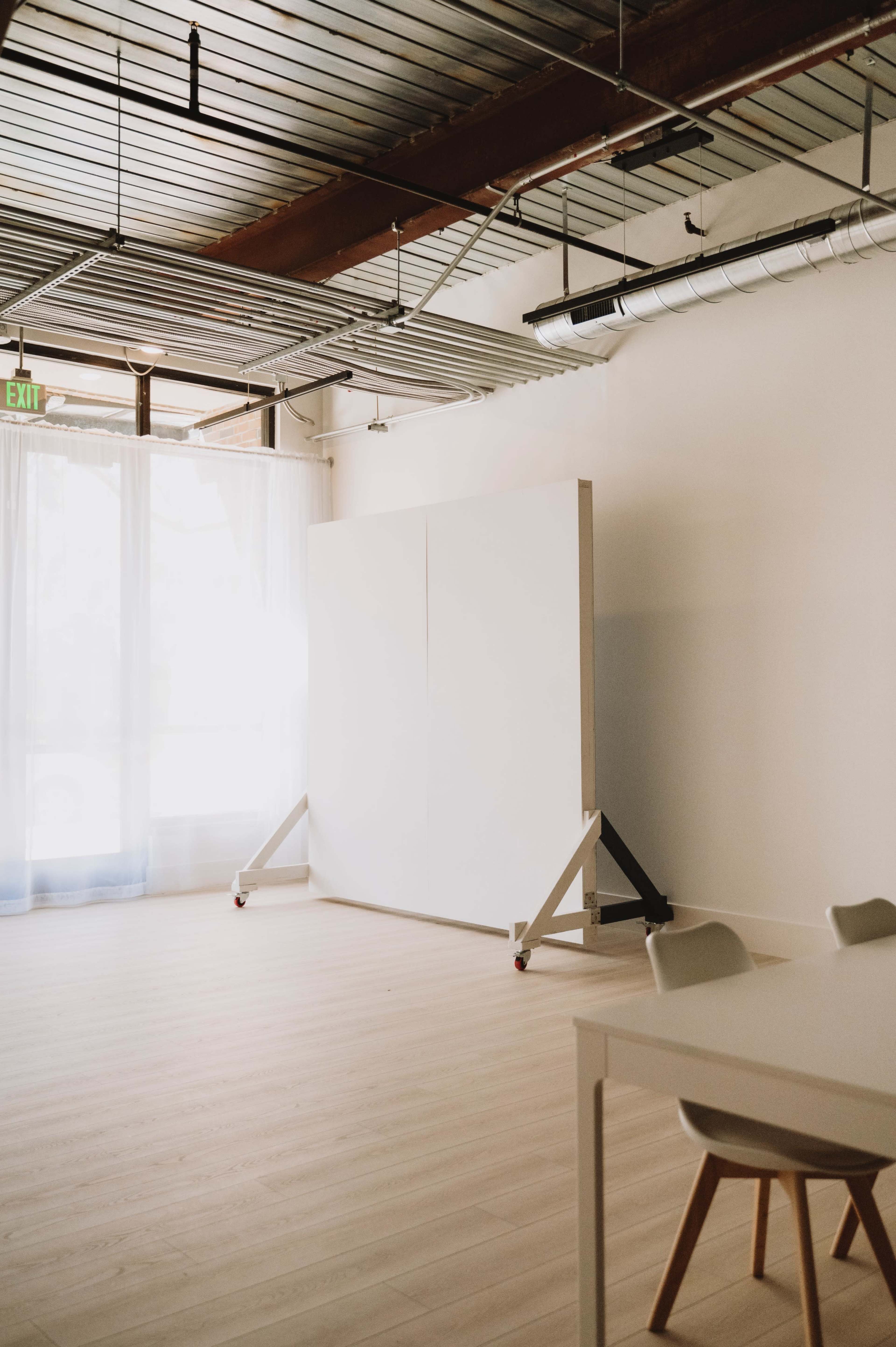 An empty room features a large, movable white panel and a simple table with chairs, illuminated by natural light through a window.