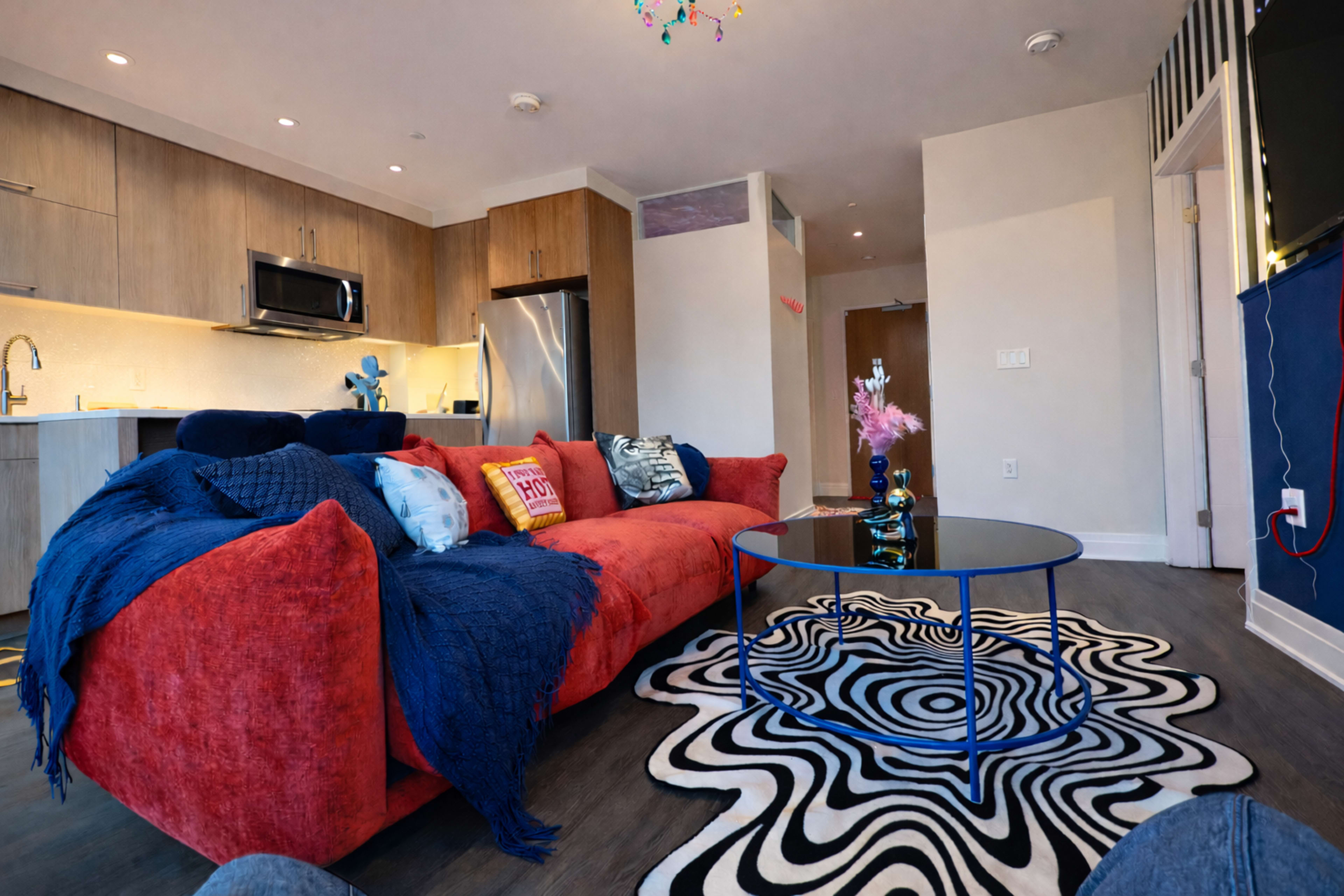 The image shows a stylish living room with a red sofa, a black and white patterned rug, a round glass coffee table, and a modern kitchen area in the background.
