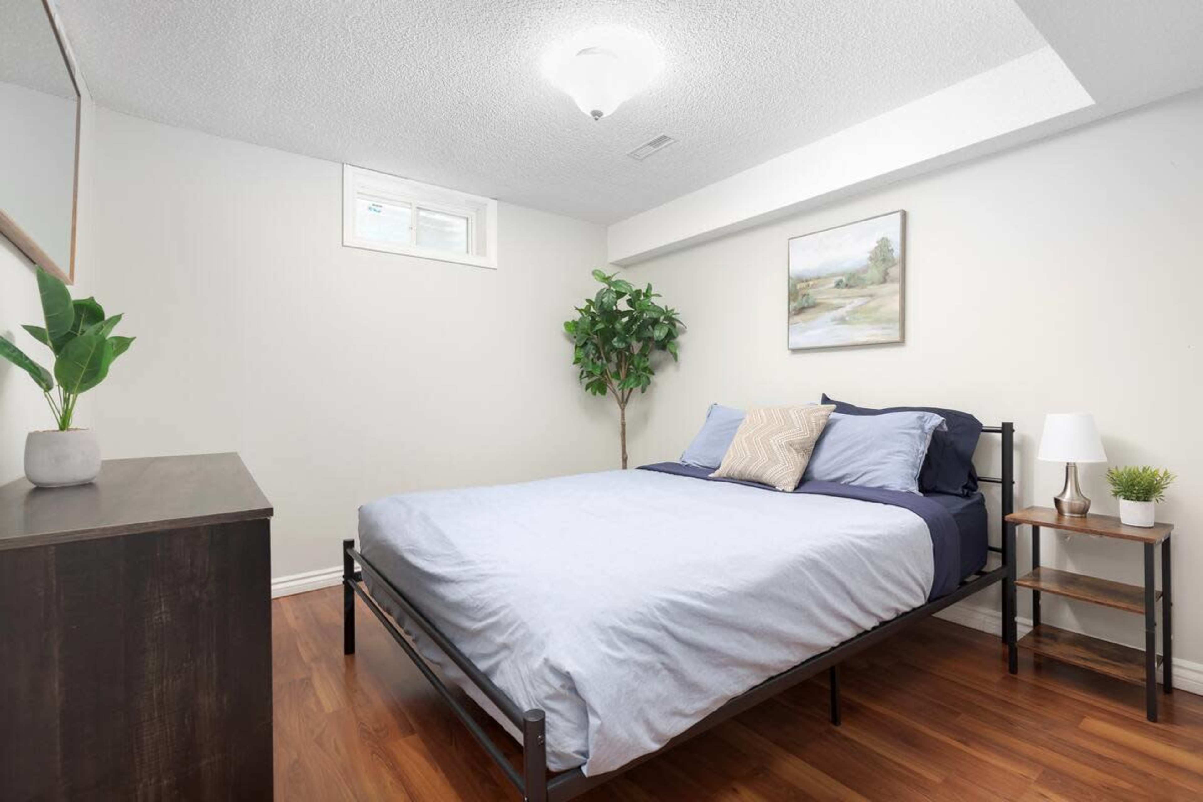 A small bedroom with a bed, a nightstand, a plant, and a dresser against a light-colored wall.