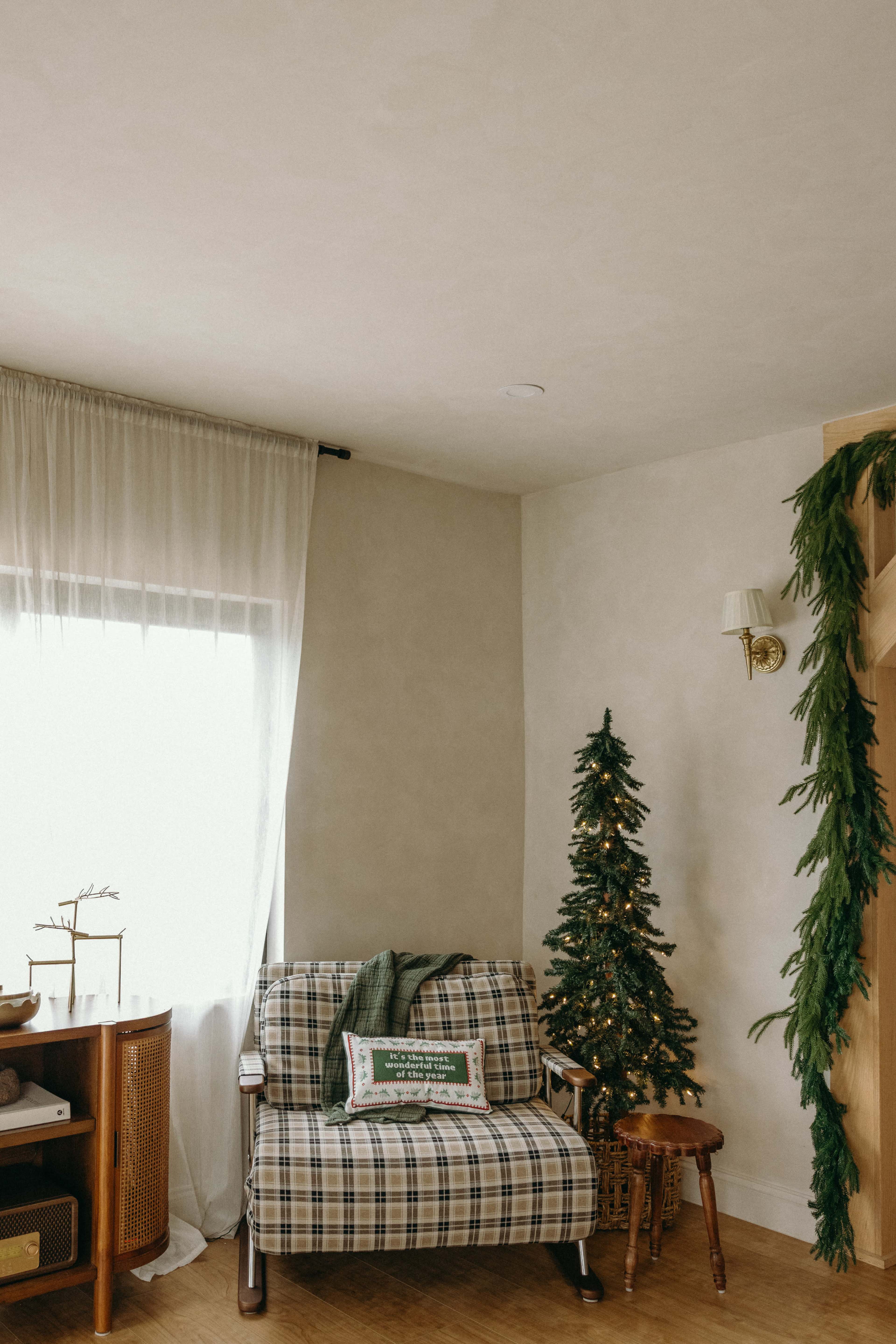 The image shows a cozy room featuring a plaid sofa, a small Christmas tree, a wooden side table, and a large window with sheer curtains.