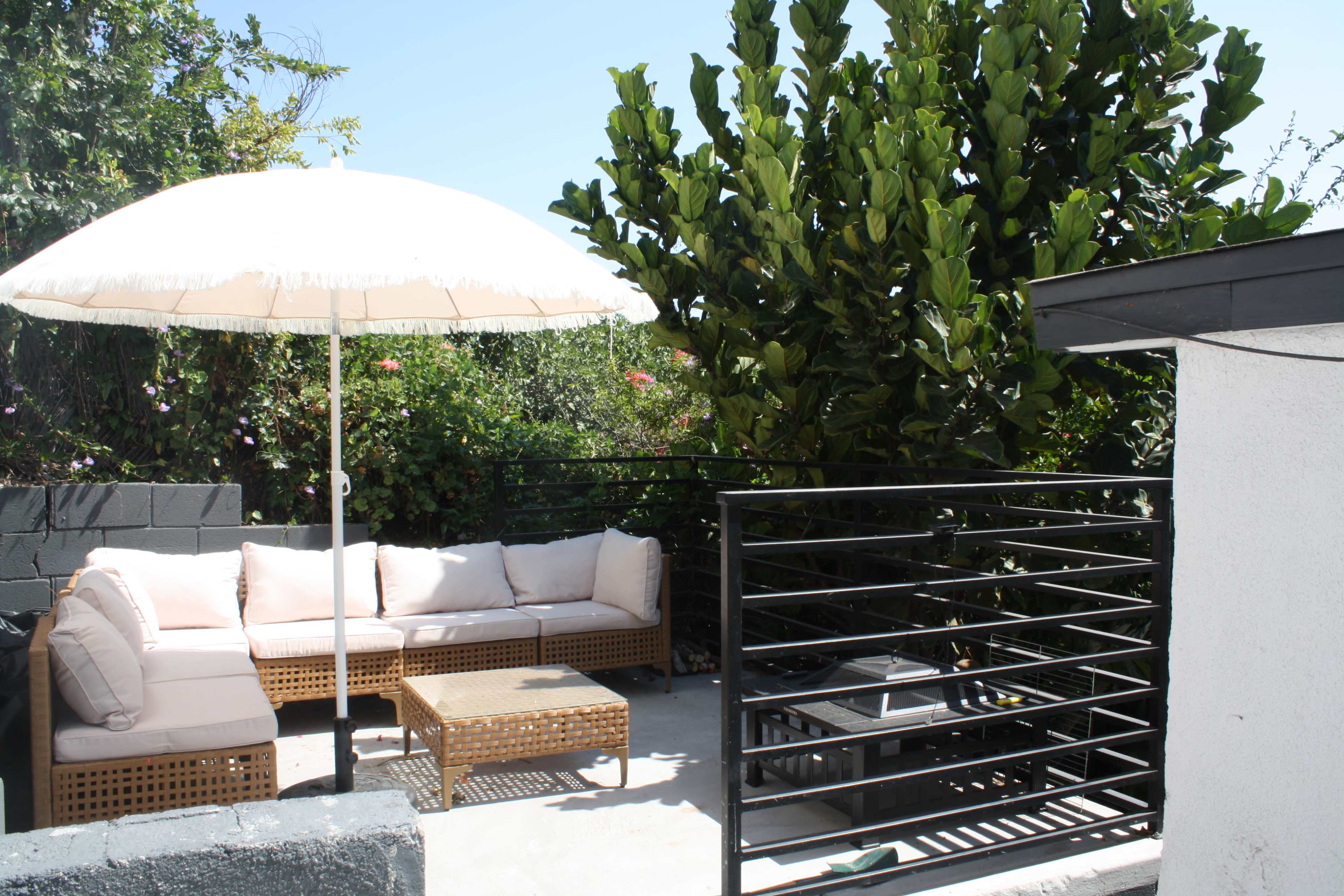 A patio features a cushioned sectional sofa and a small table under a large umbrella, surrounded by greenery.