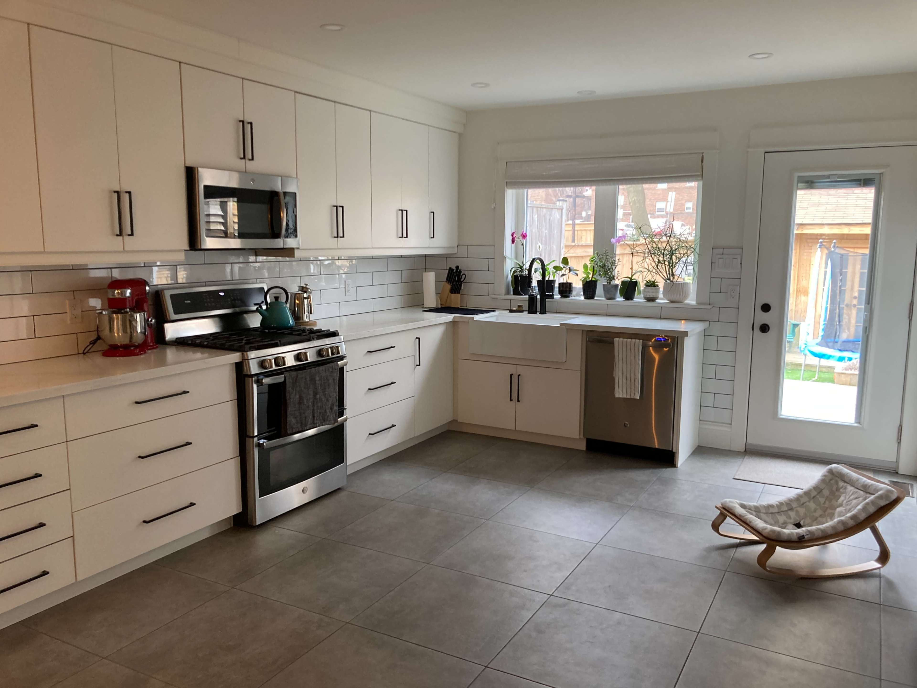 A modern kitchen features white cabinets, stainless steel appliances, and a large window with plants on the sill.