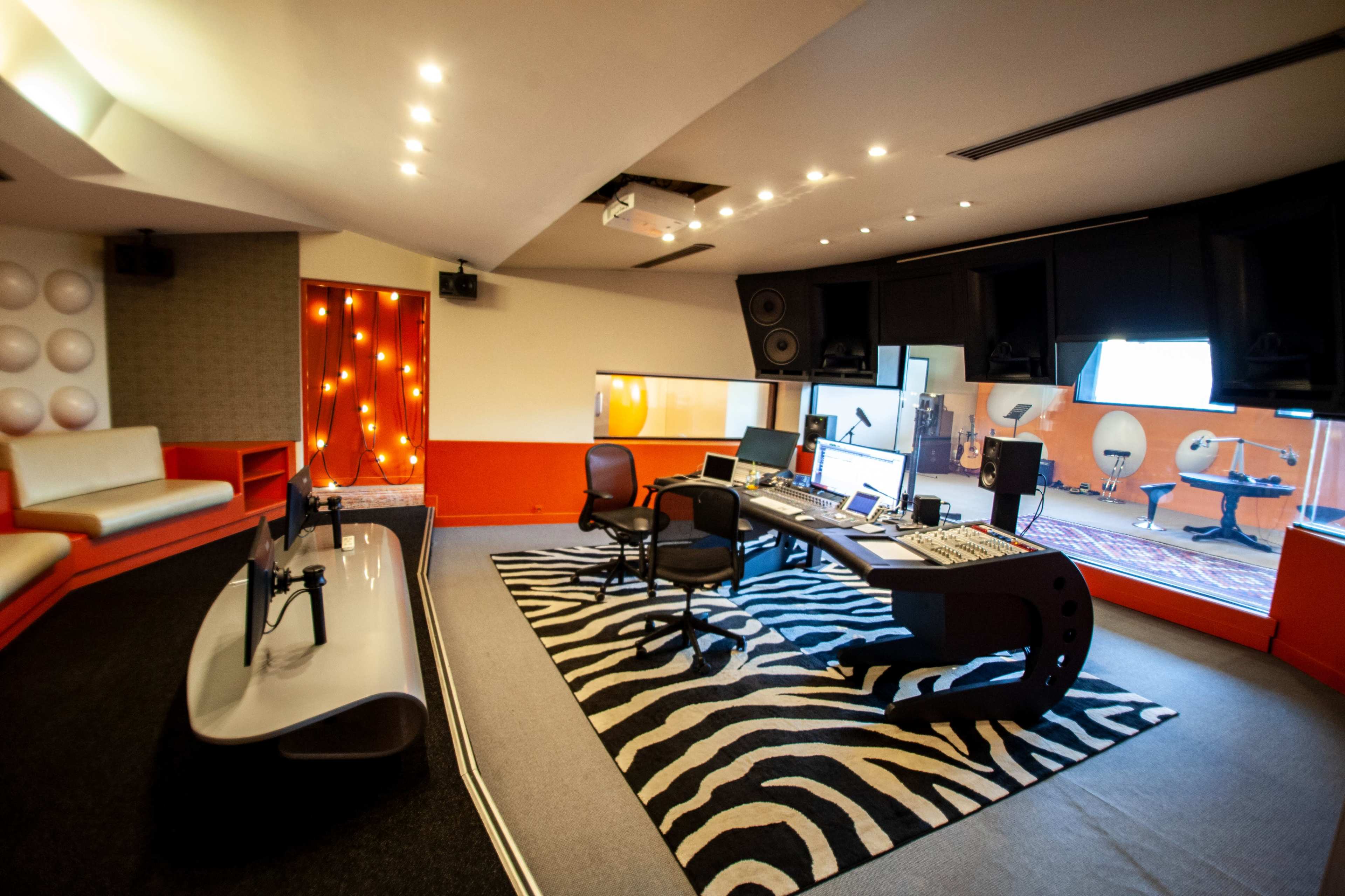A modern music studio featuring a large mixing desk, computer equipment, and soundproof walls, with instruments visible through a glass wall.