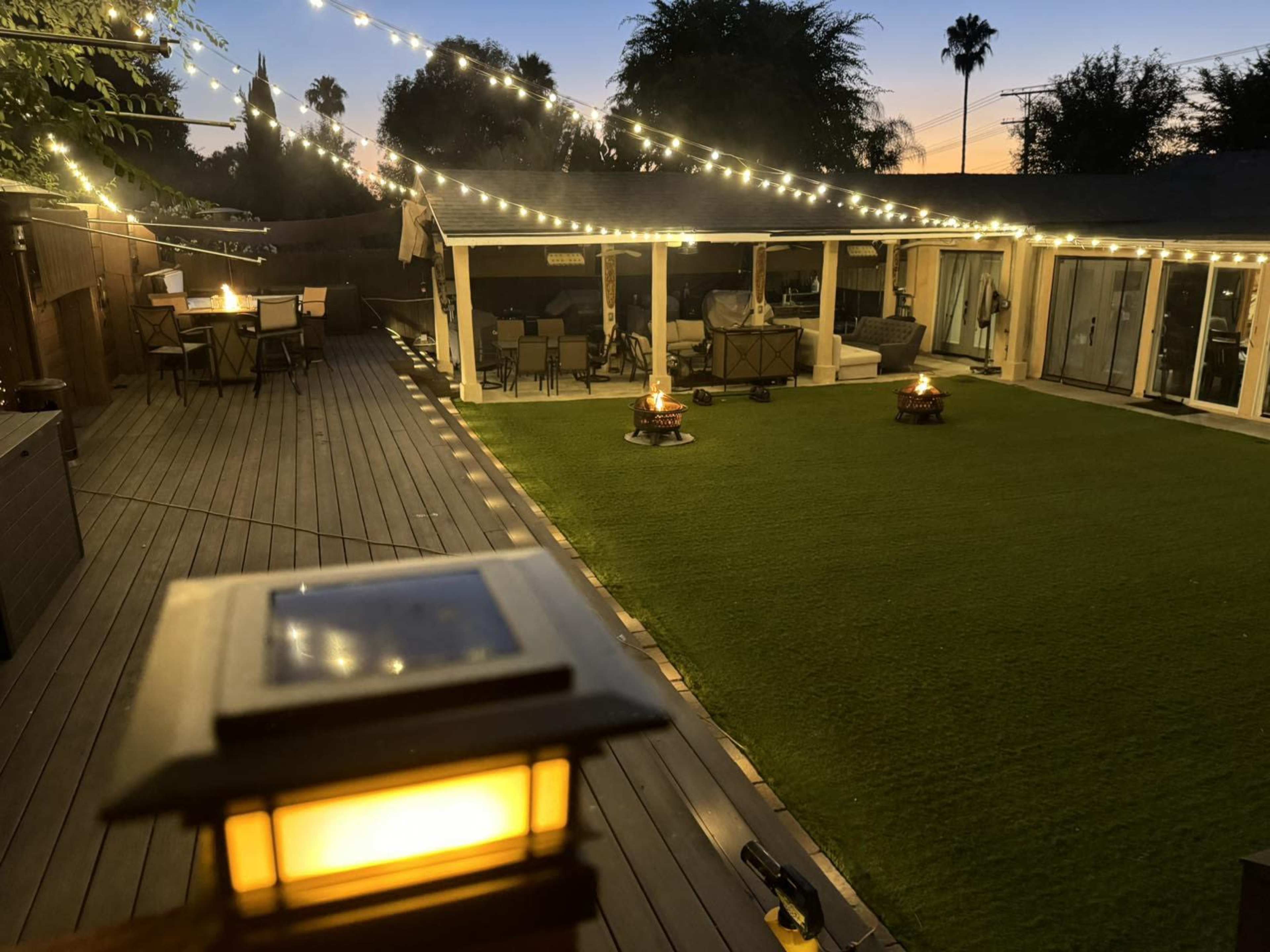 A well-lit outdoor space features string lights above a grassy area with seating and a dining table, surrounded by palm trees and a wooden deck.