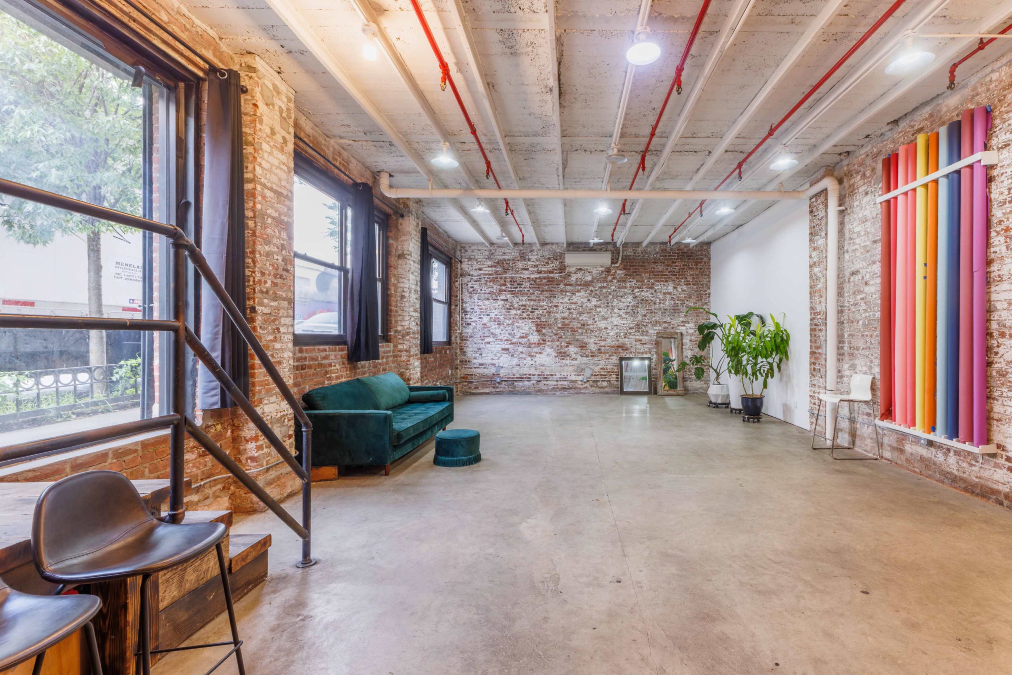 The image shows a spacious, modern room with exposed brick walls, concrete flooring, and colorful vertical stripes decorating one wall.