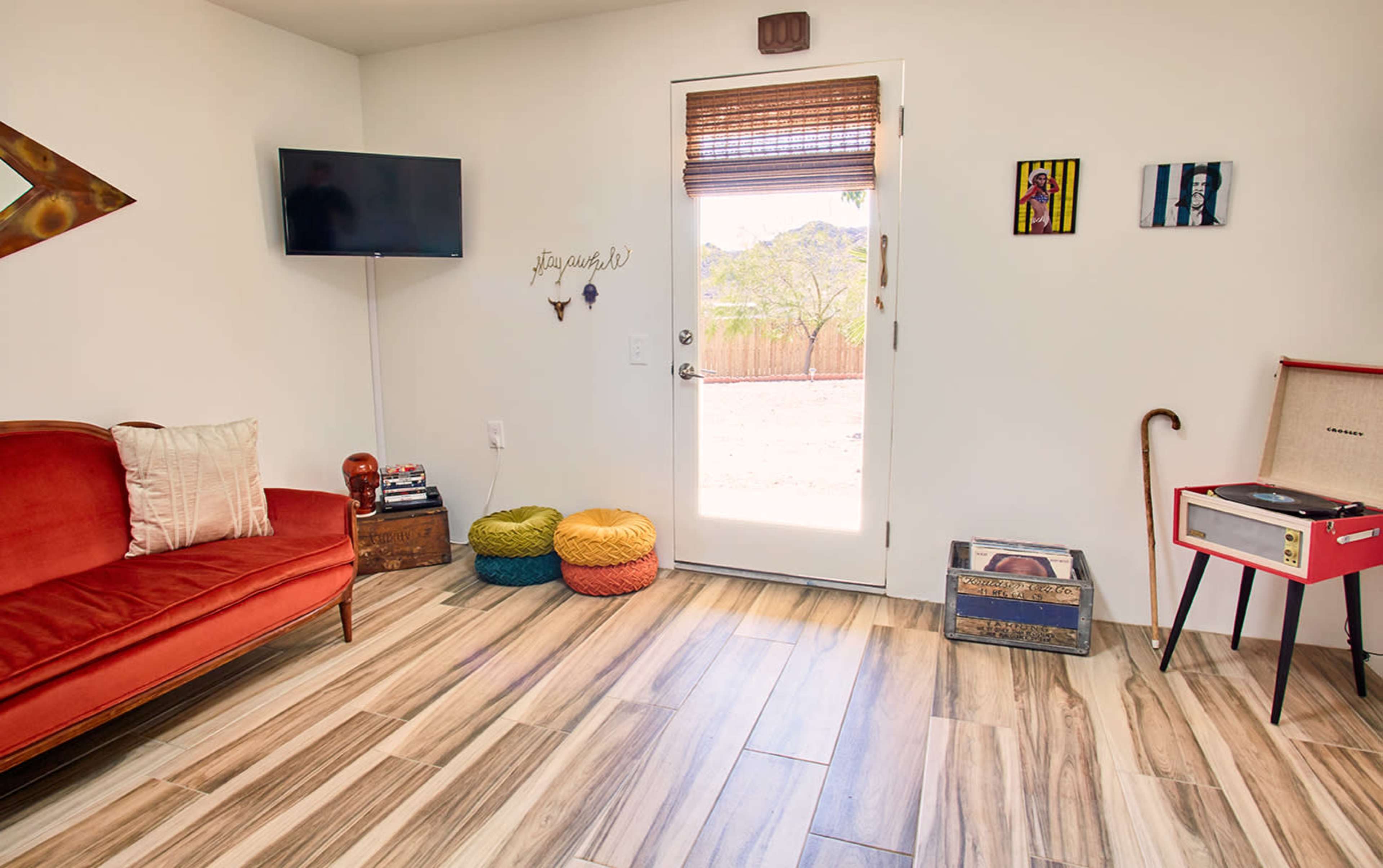 The image shows a cozy interior space featuring a red couch, a wall-mounted television, and colorful cushioned seating near a door leading to an outdoor view.