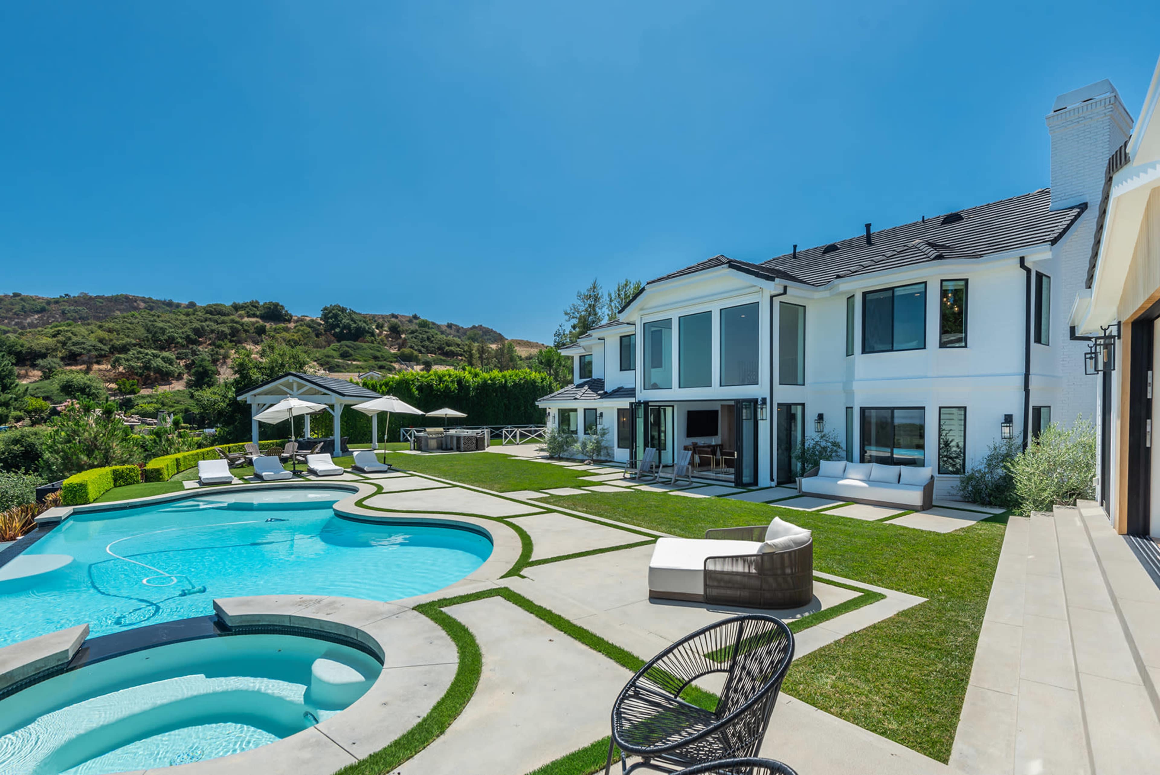 A modern house with large windows overlooks a swimming pool and landscaped yard in a sunny setting.