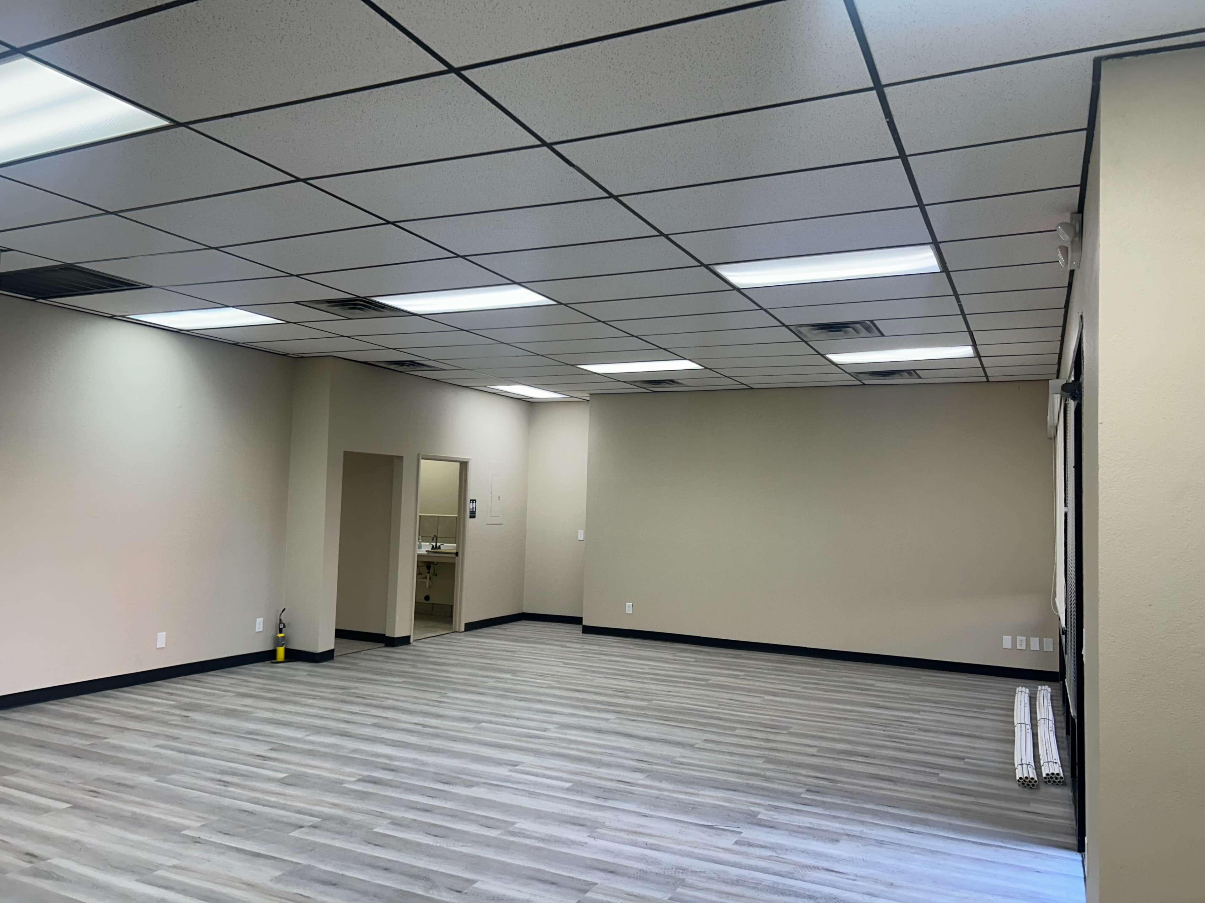 The image shows an empty room with light gray flooring, plain beige walls, and fluorescent ceiling lights.