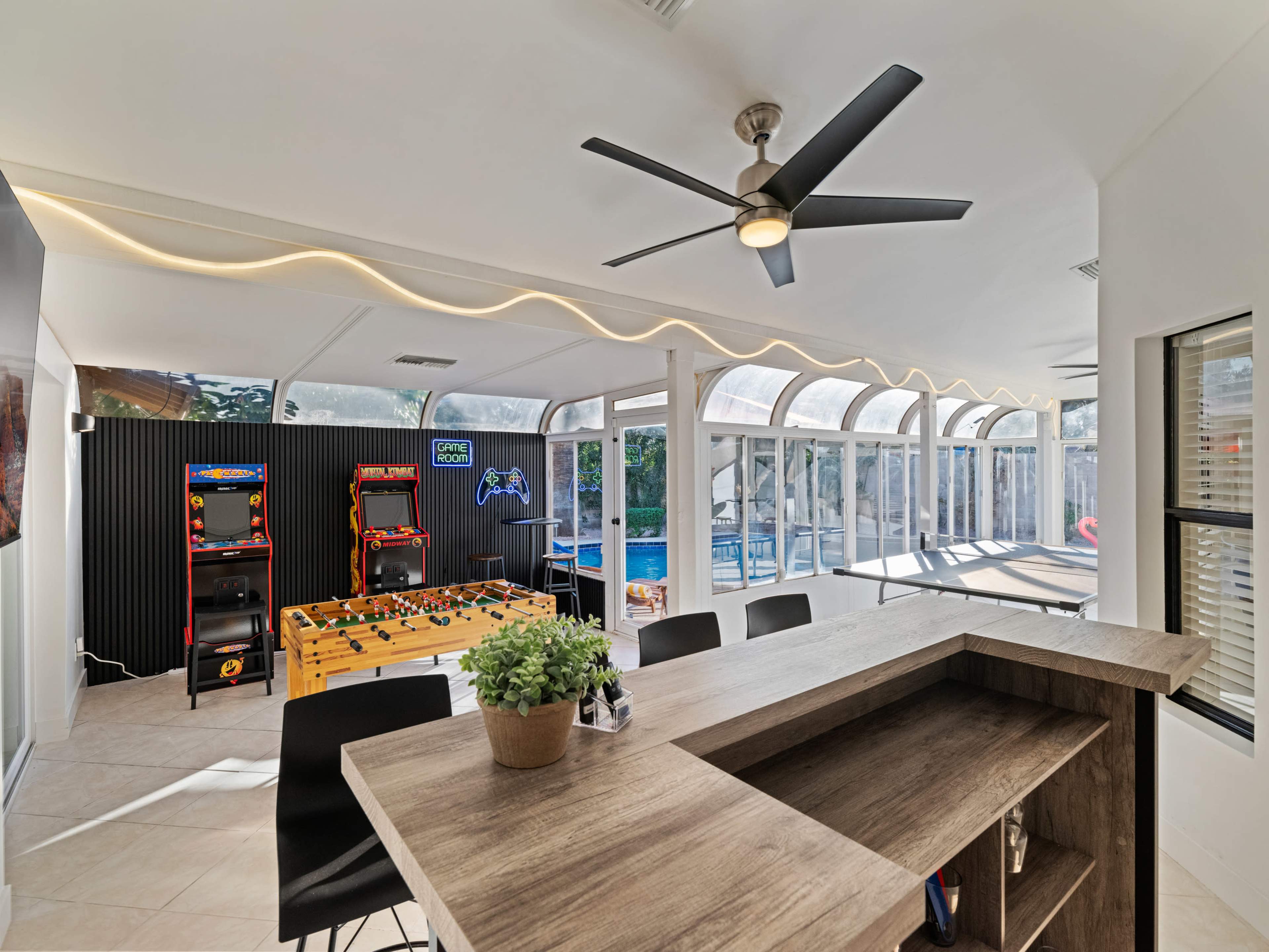 A modern game room featuring arcade machines, a foosball table, and a view of a pool through large windows.