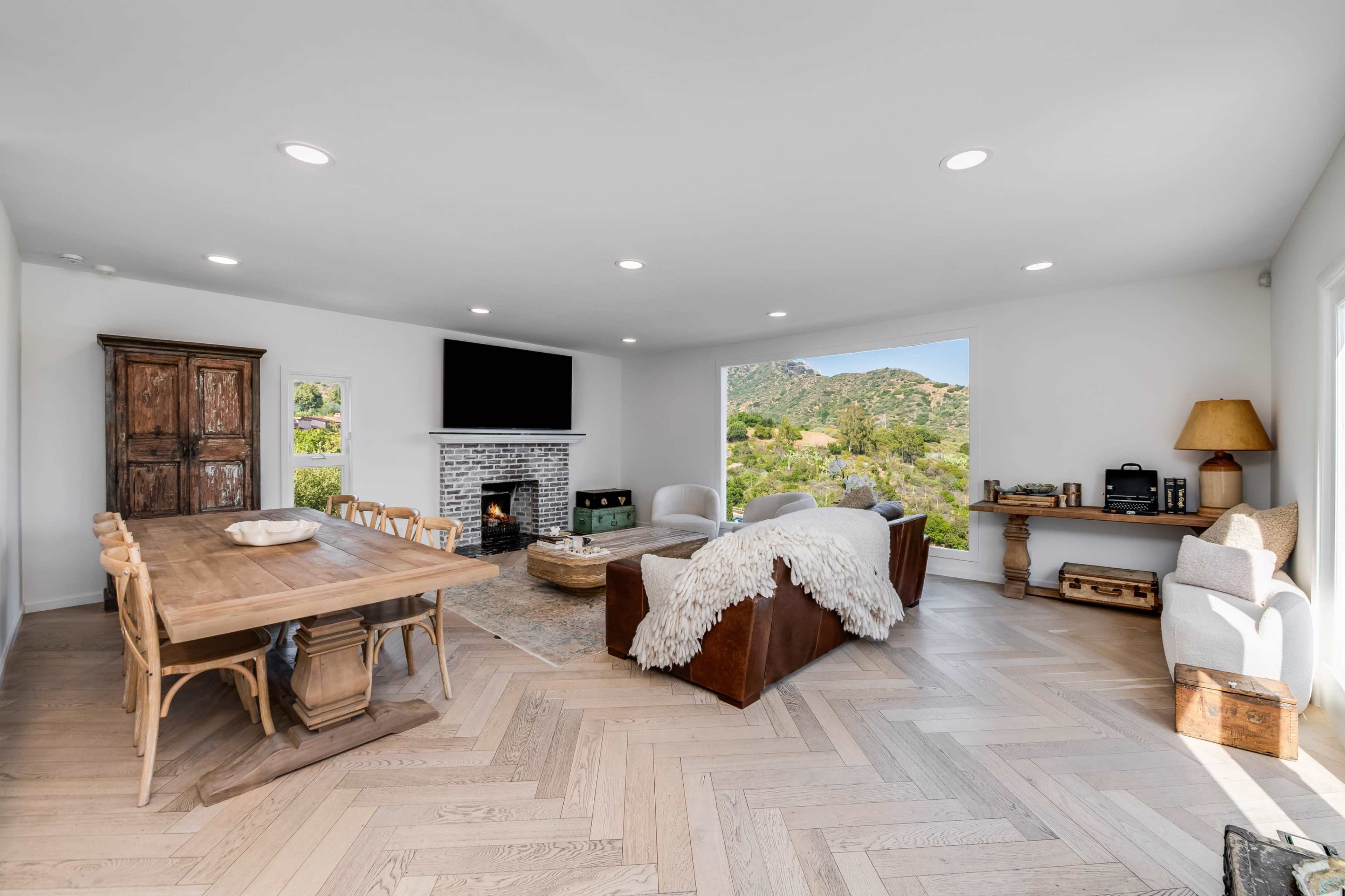 A modern living and dining area features a large window with a view of greenery, a cozy sofa, a dining table with chairs, and a fireplace.