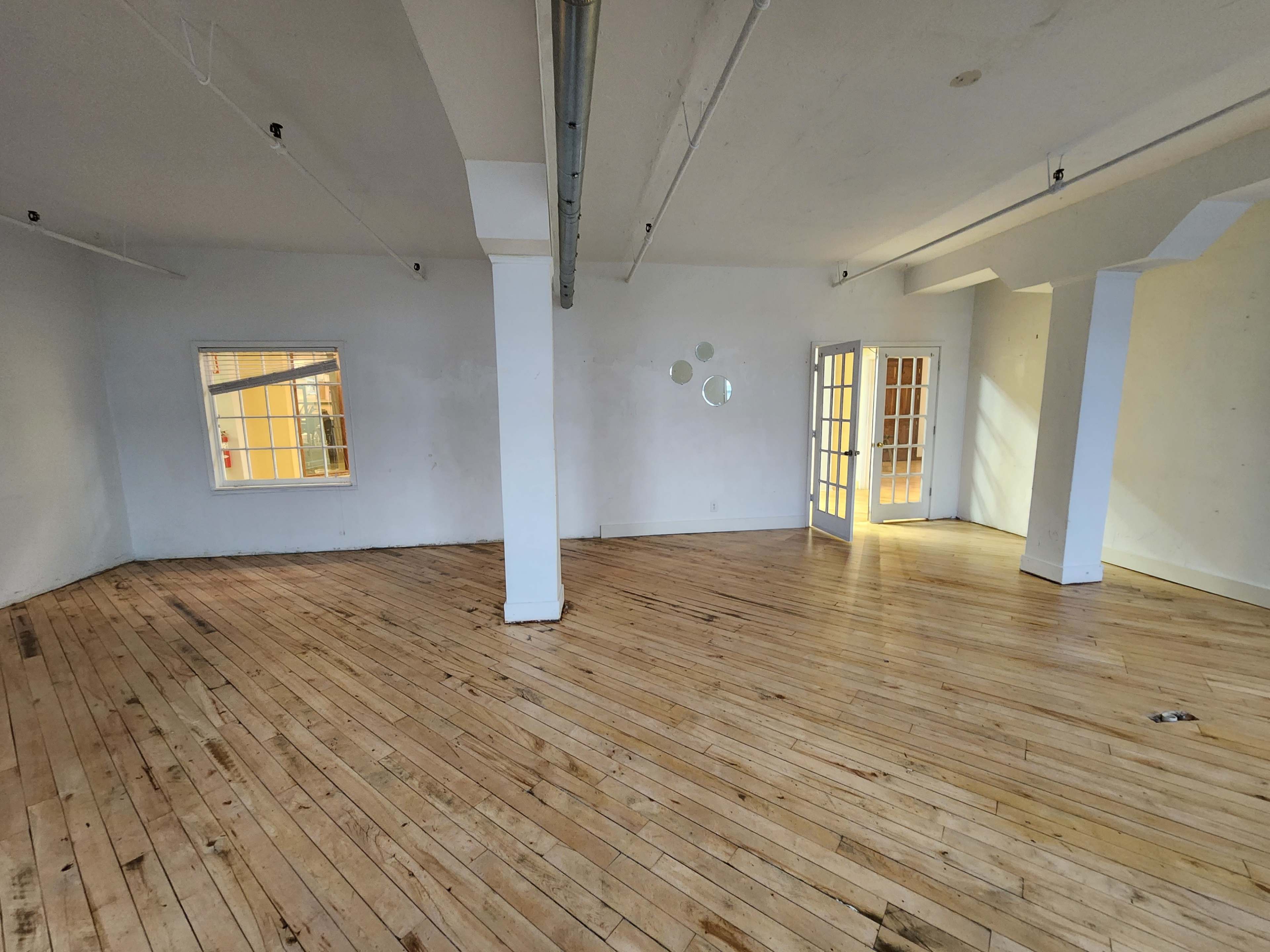 An empty room with wooden floors, white walls, large windows, and a set of glass-paneled doors leading to another space.