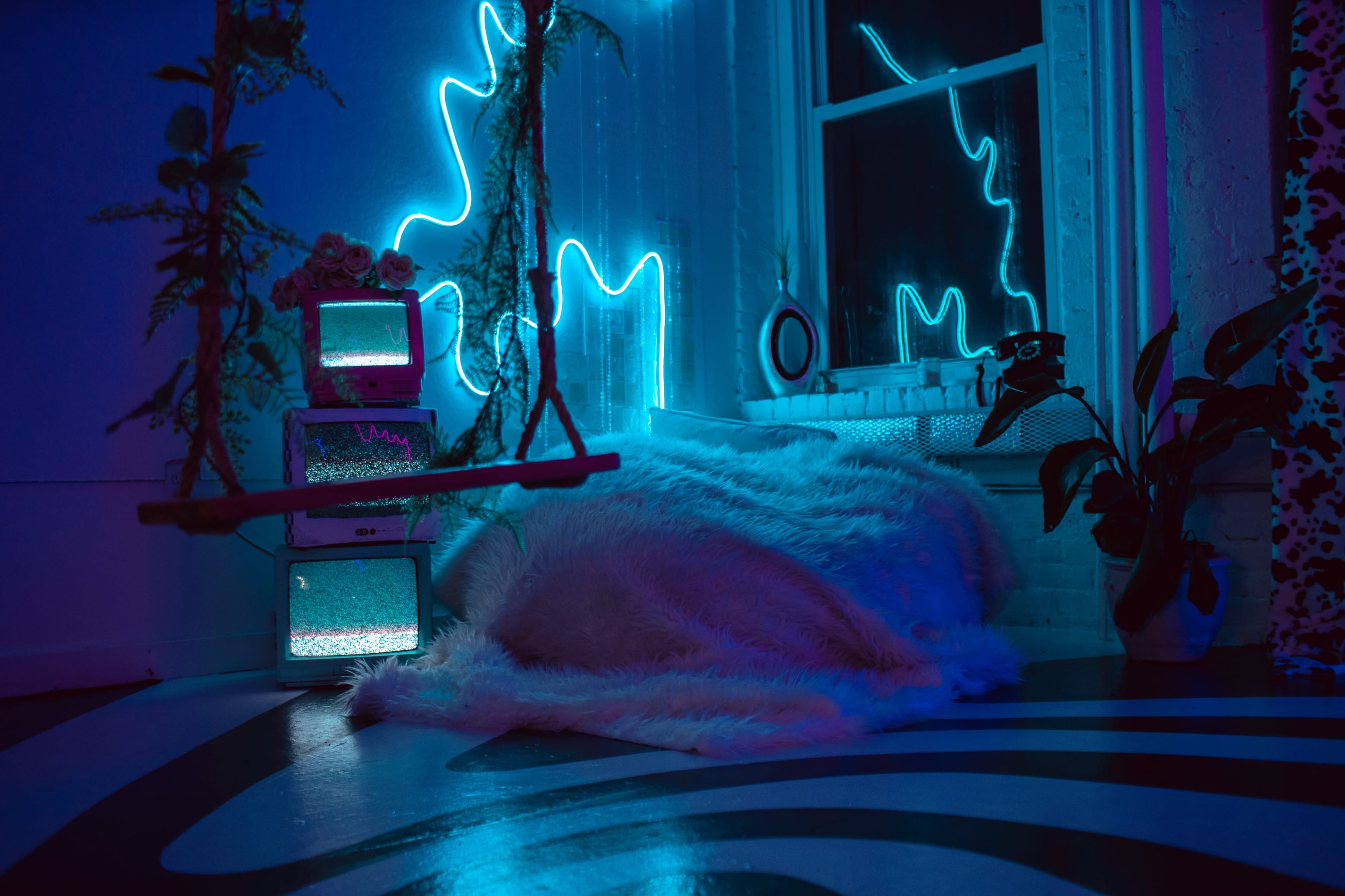A cozy room features a swing, glowing neon lights, a plush rug, and vintage televisions beside a large window.