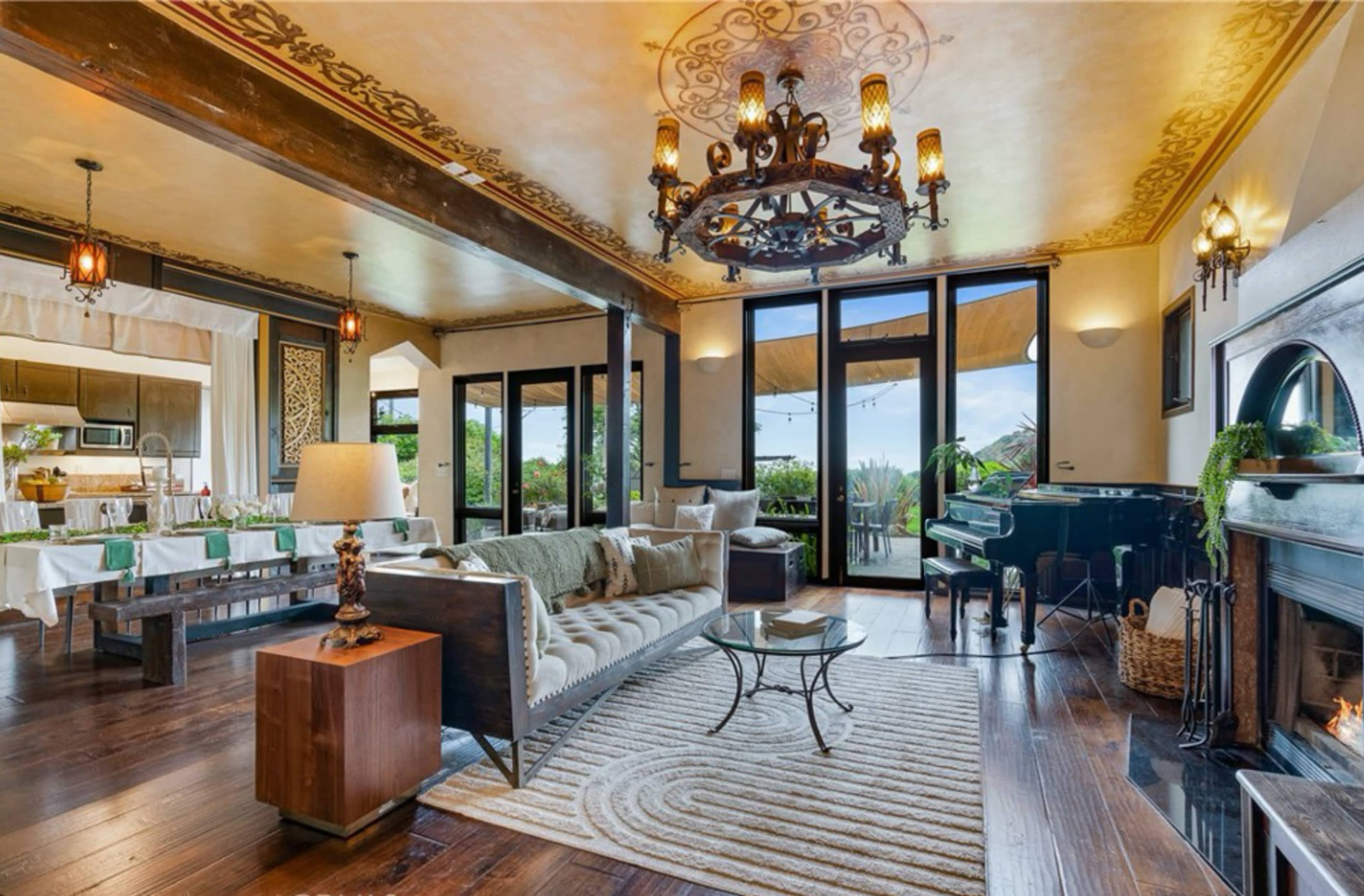 The image shows a warmly decorated living room featuring a chandelier, a grand piano, and large glass doors opening to a patio, with a cozy sofa and wooden furniture.