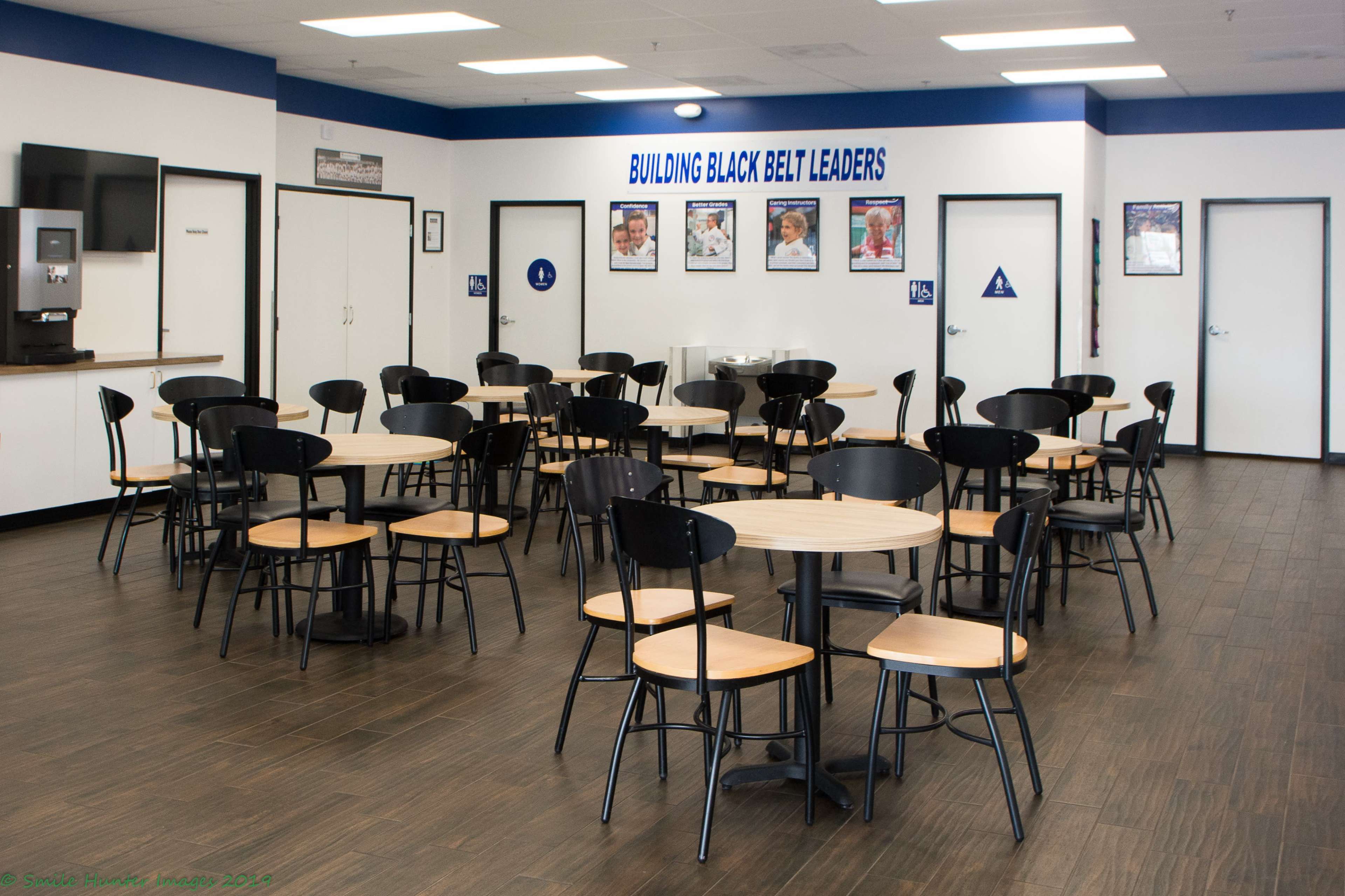 A spacious, well-lit dining area with multiple round tables and black chairs, featuring motivational posters on the walls.