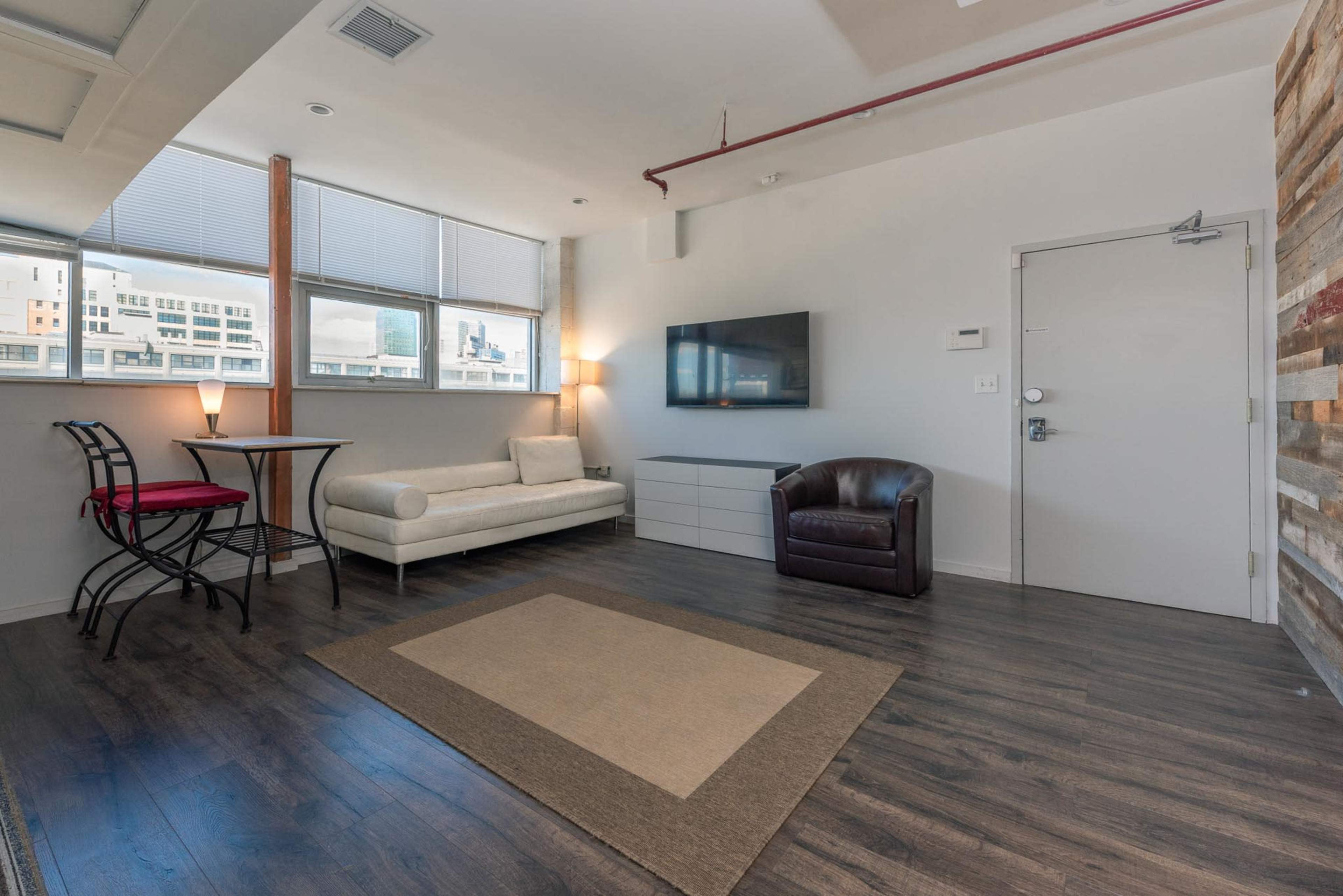 A modern living room with a white sofa, a brown armchair, a small dining table, a television on the wall, and large windows providing natural light.