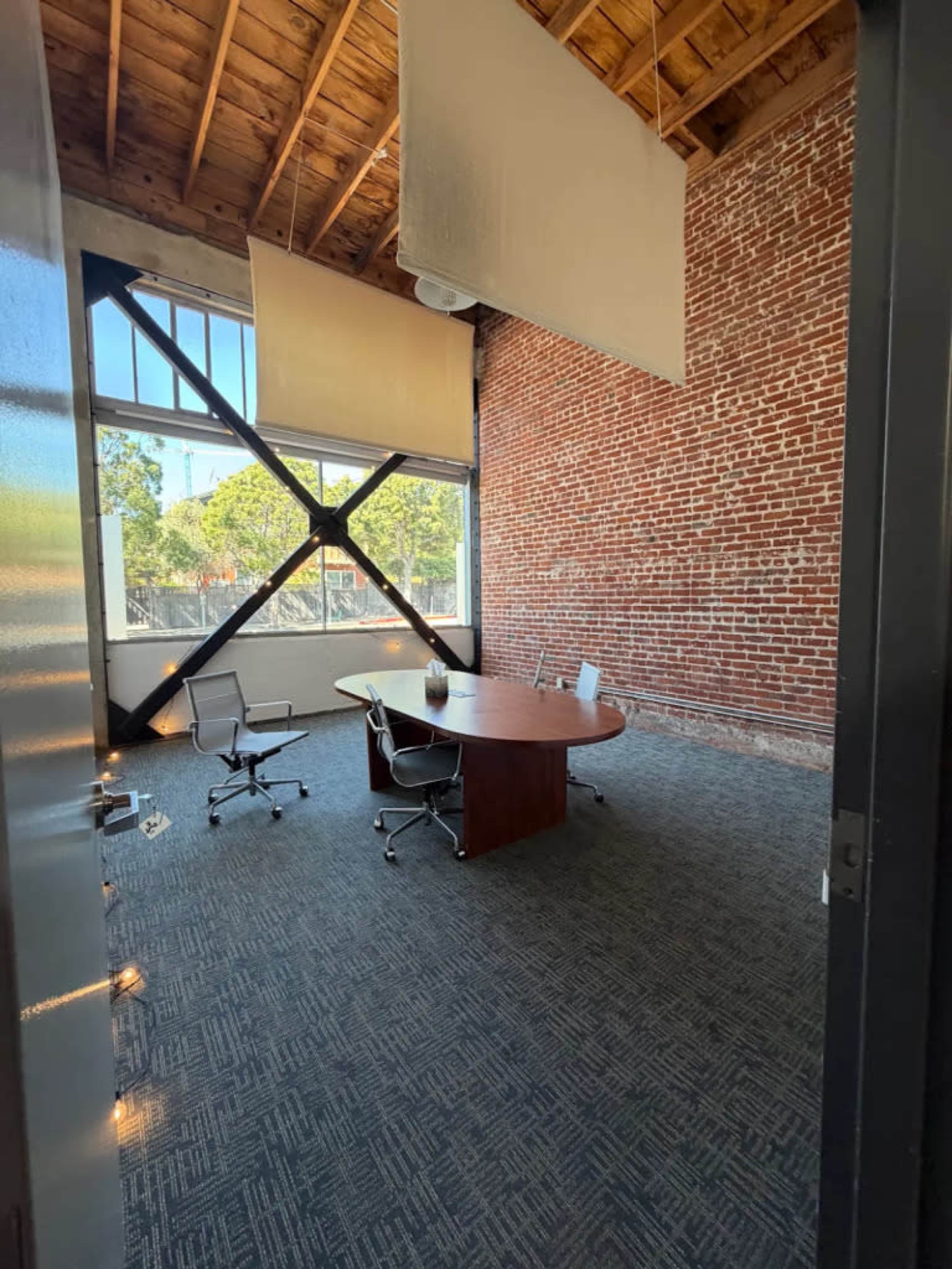 The image shows a modern conference room with a round wooden table and several ergonomic chairs, situated against a backdrop of exposed brick walls and a large window with natural light.