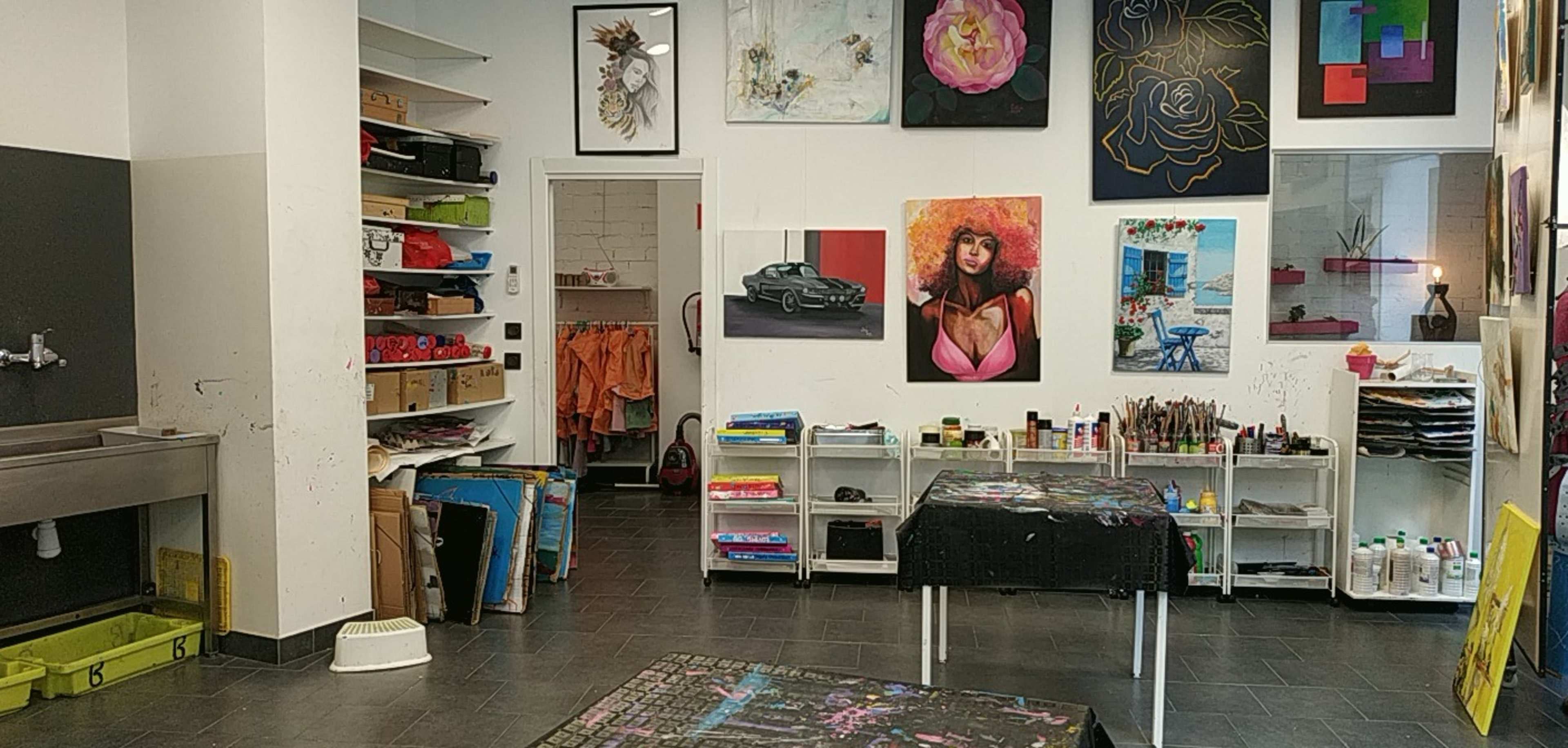 The image shows an art studio with various paintings displayed on the walls, shelves filled with art supplies, and work tables in the center.