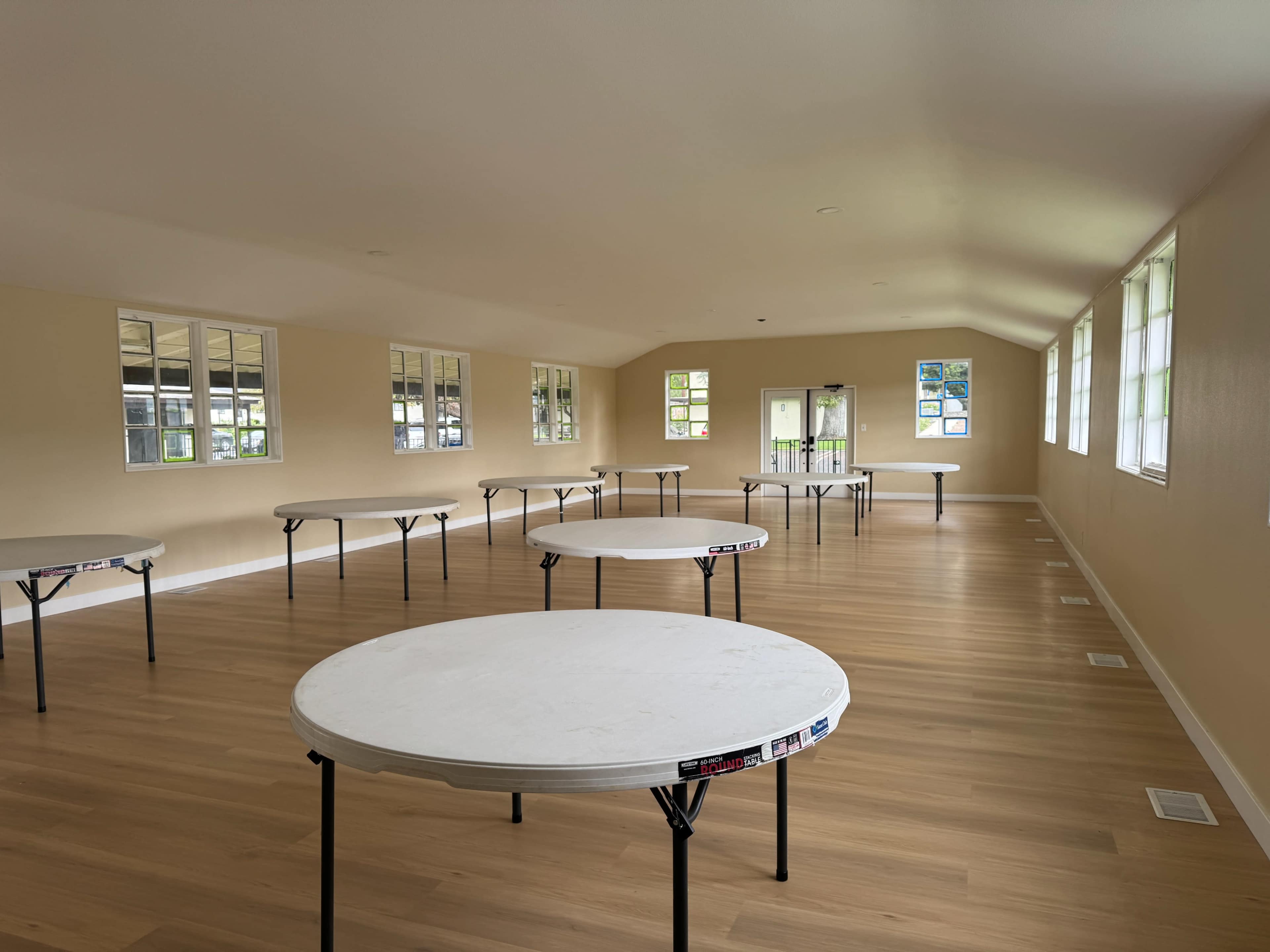 The image shows a spacious indoor area with several round tables arranged around the room and large windows letting in natural light.
