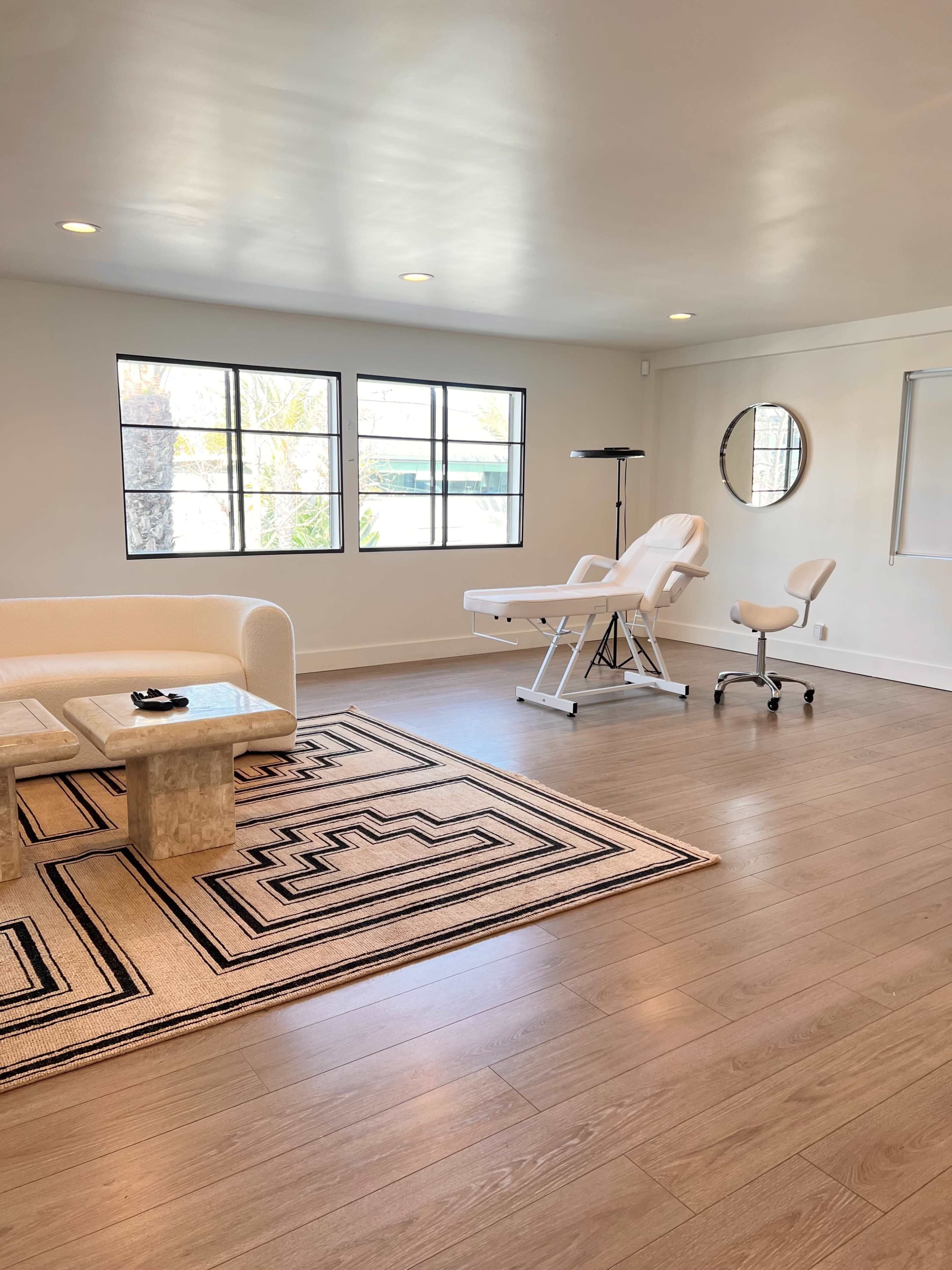 A bright, minimalist treatment room featuring a white couch, a marble coffee table, and a treatment chair beside a large window.