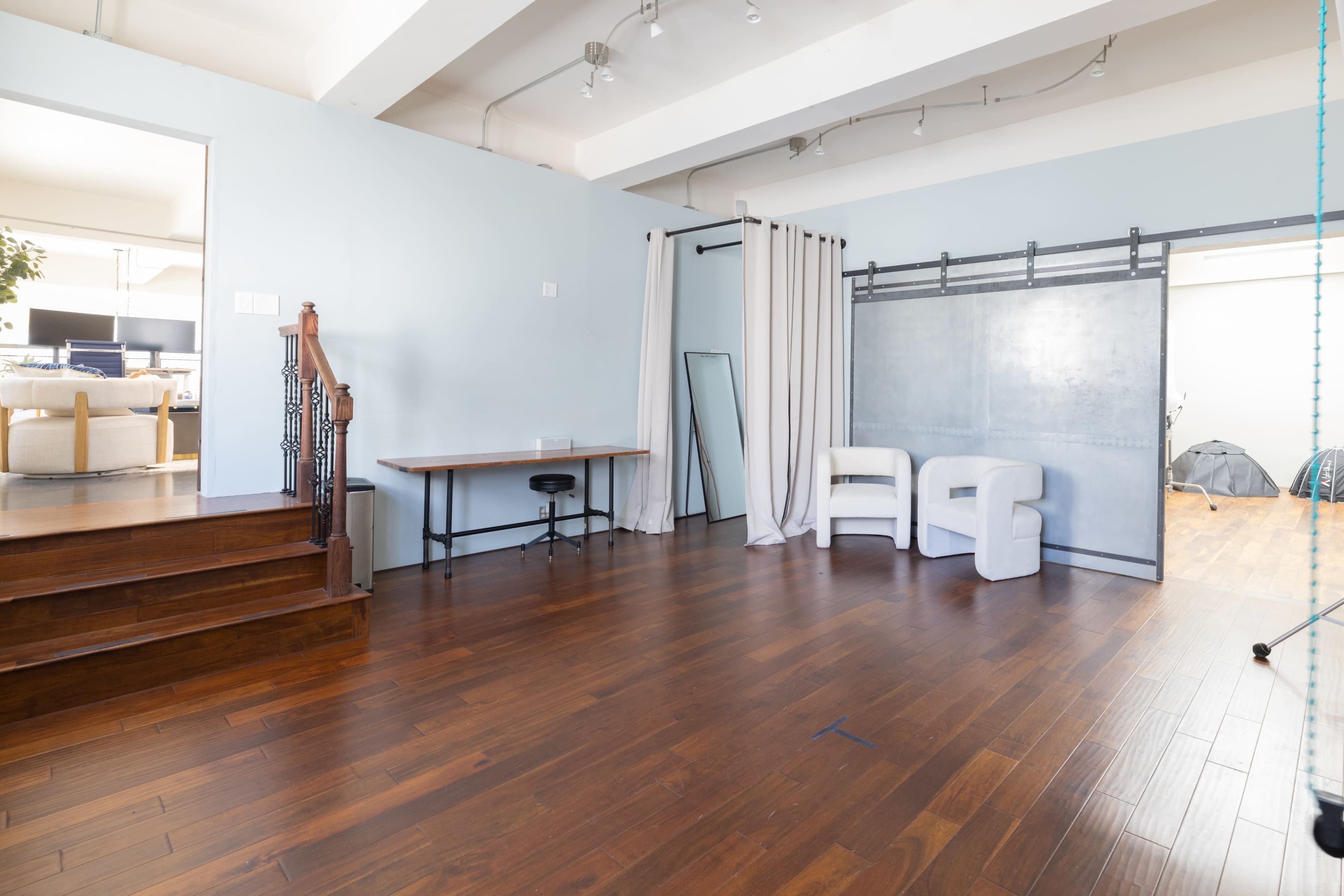 The room features hardwood flooring, a table against the wall, a mirror, a set of white chairs, and a partition next to a staircase.