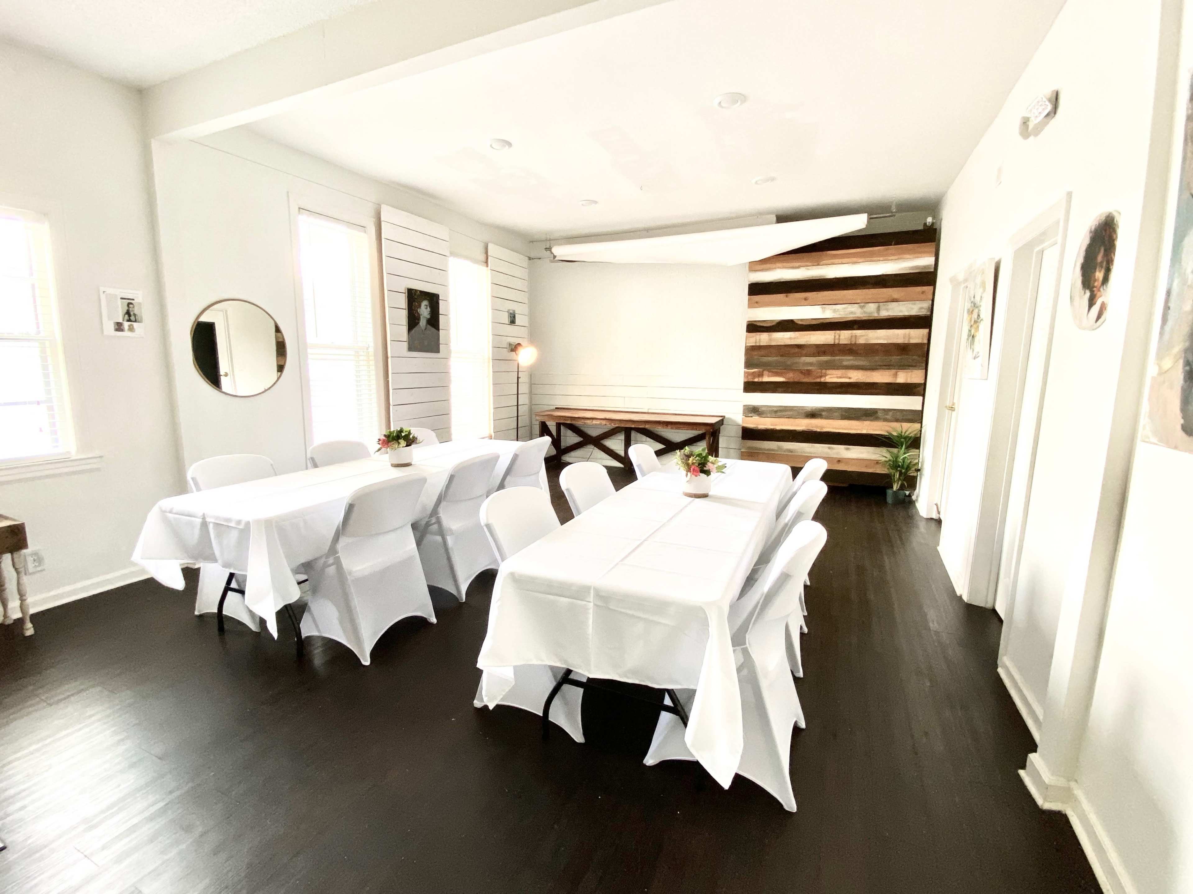 A bright, modern room features two long tables covered with white tablecloths and surrounded by folding chairs, against a backdrop of wooden accent walls.