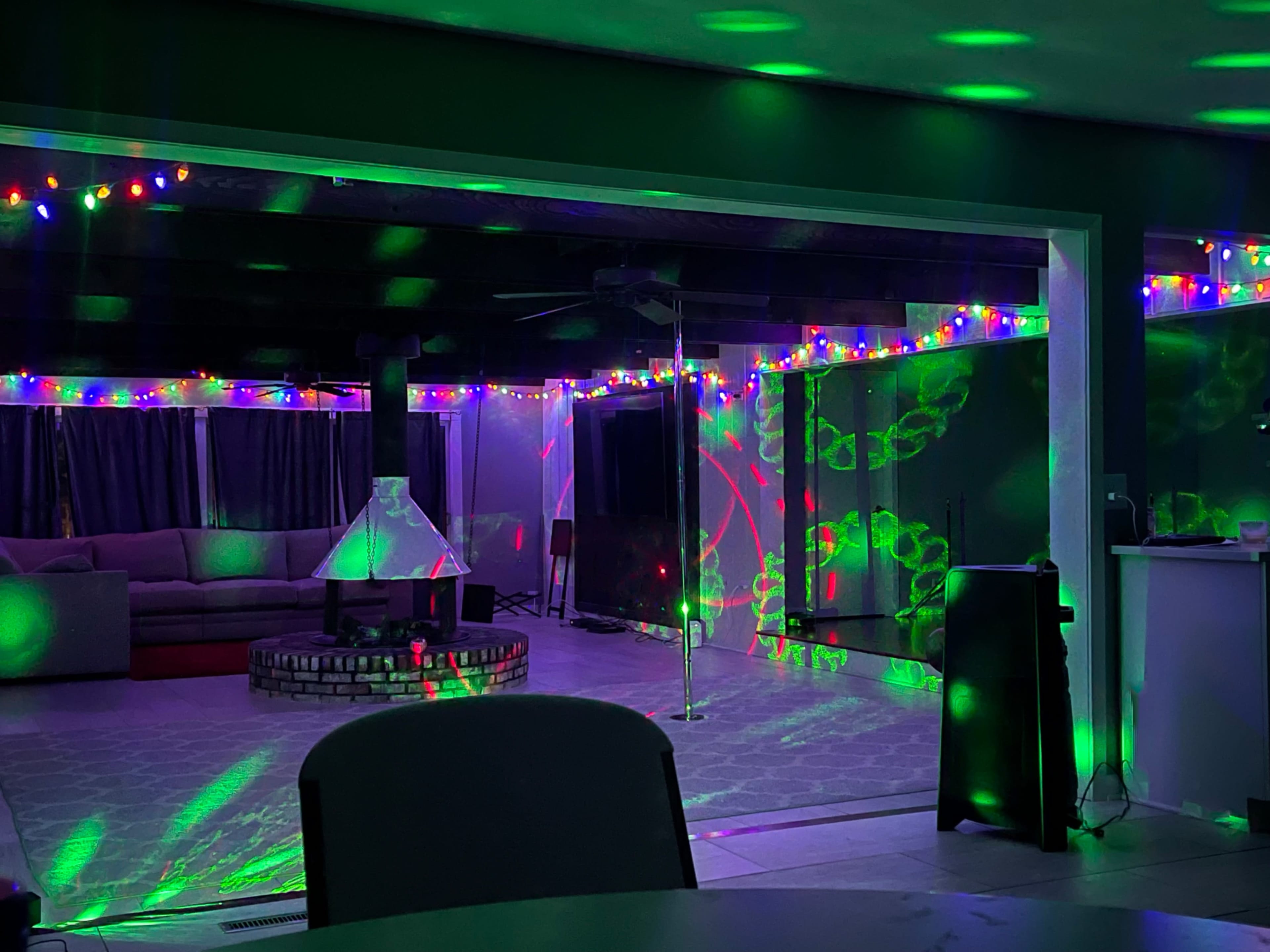 A spacious indoor area is decorated with colorful lights, featuring a seating arrangement, a central firepit, and a dance pole.