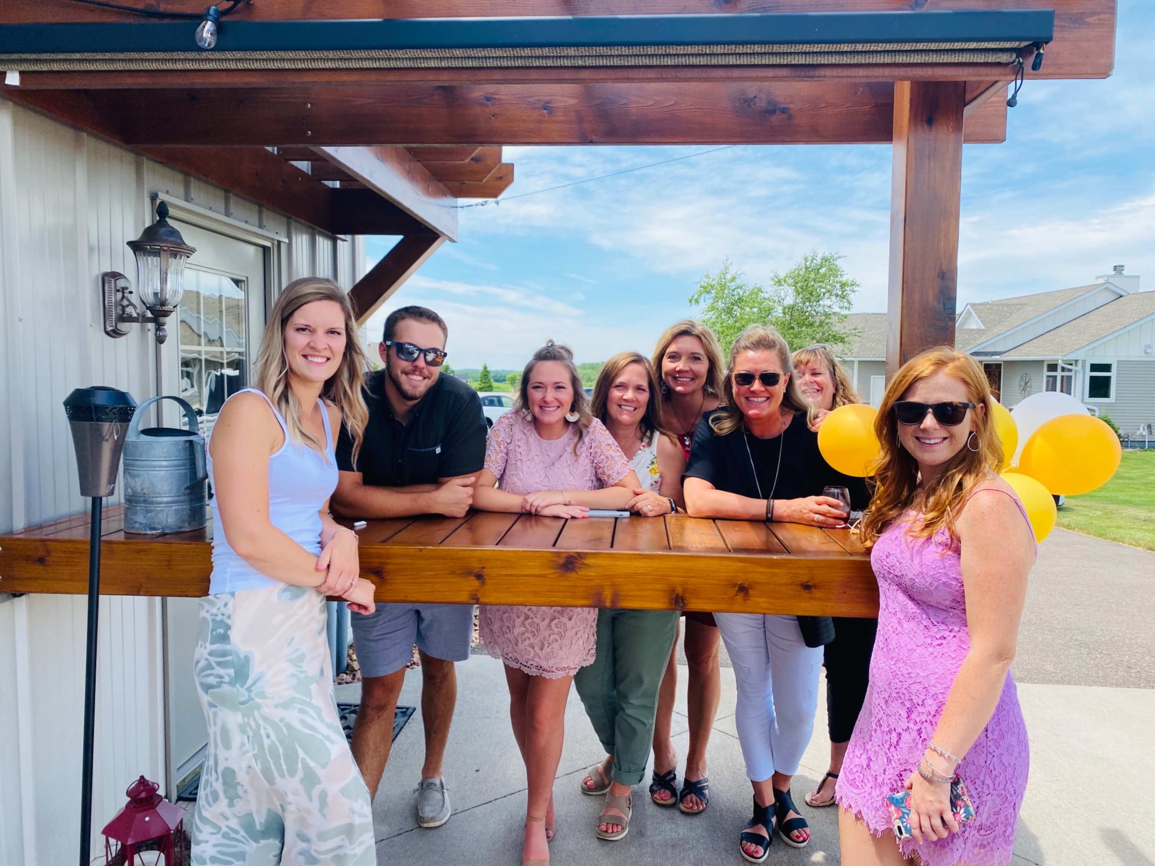 A group of eight people poses together at an outdoor gathering near a wooden bar, with balloons and a clear blue sky in the background.