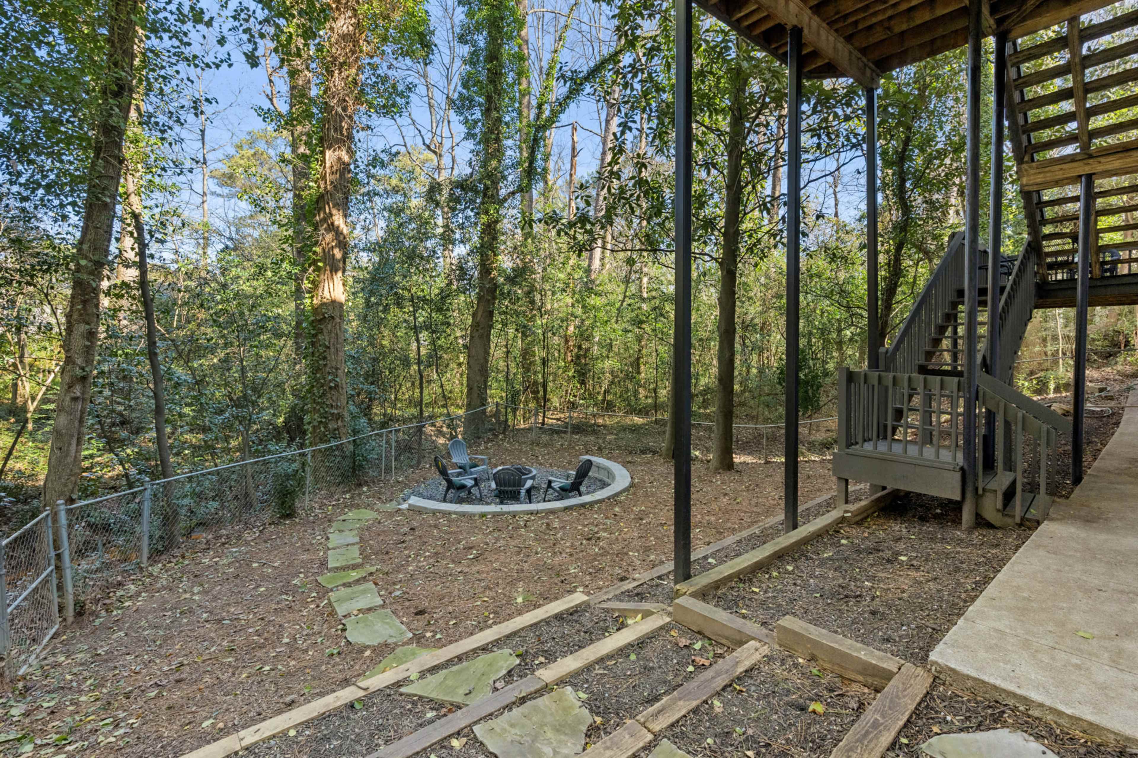 The image shows a backyard featuring a stone fire pit with seating, surrounded by trees and a fenced area, alongside a staircase leading to an elevated deck.
