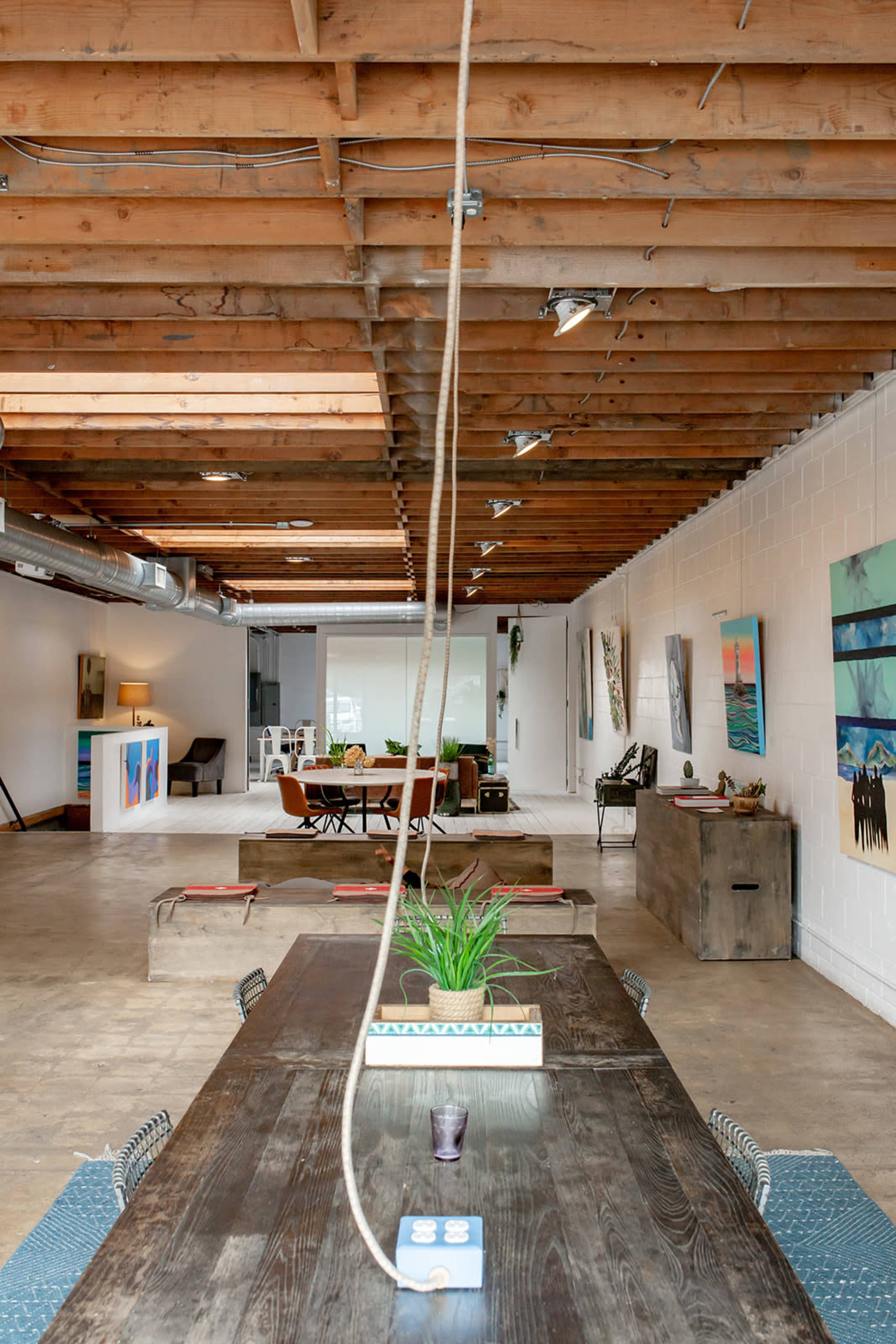 The image shows a spacious, industrial-style interior with a long wooden table and various artwork displayed on the walls.