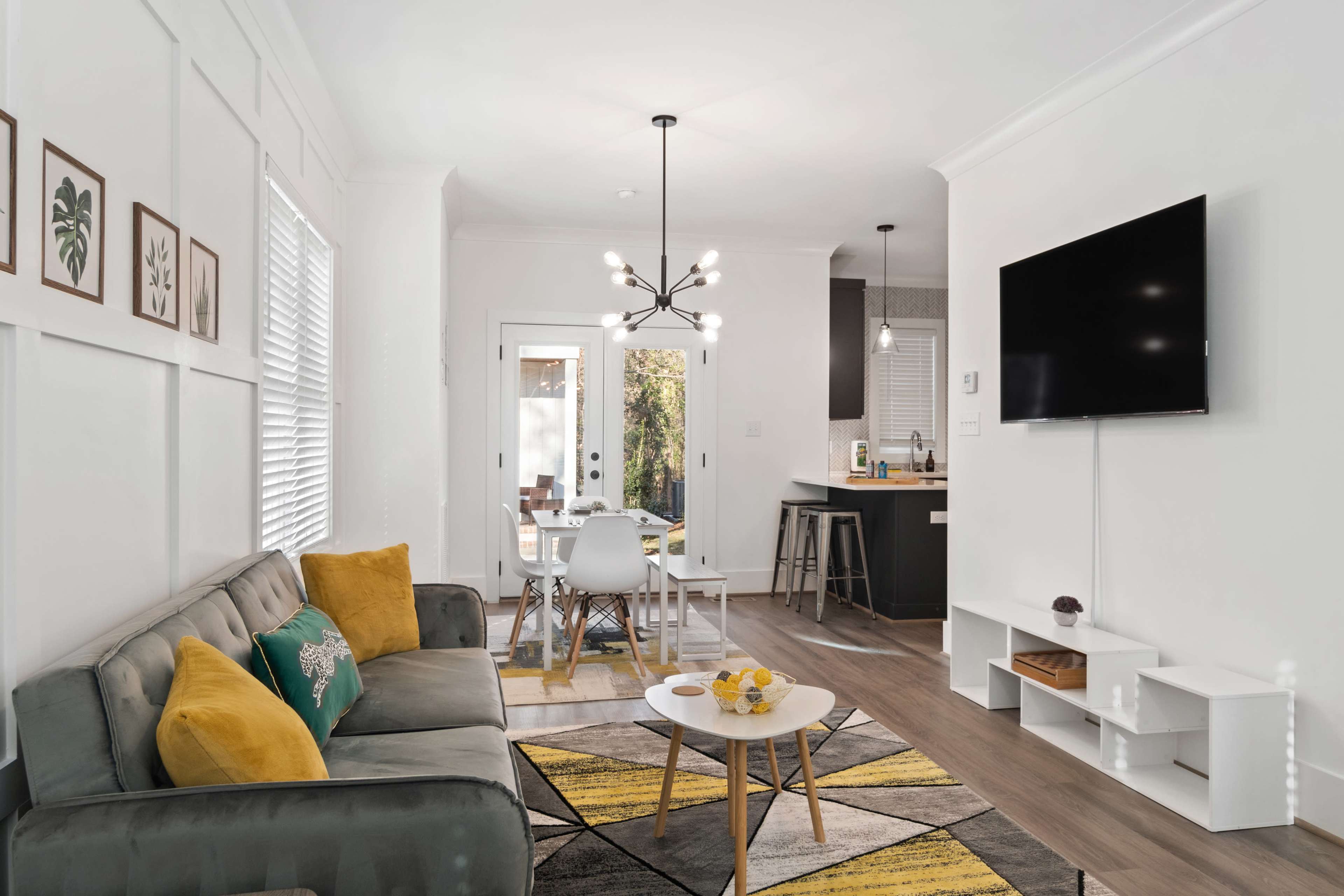 A modern living room features a gray sofa with yellow accent pillows, a round coffee table, and a wall-mounted television, leading to a dining area with a glass door opening to an outdoor space.