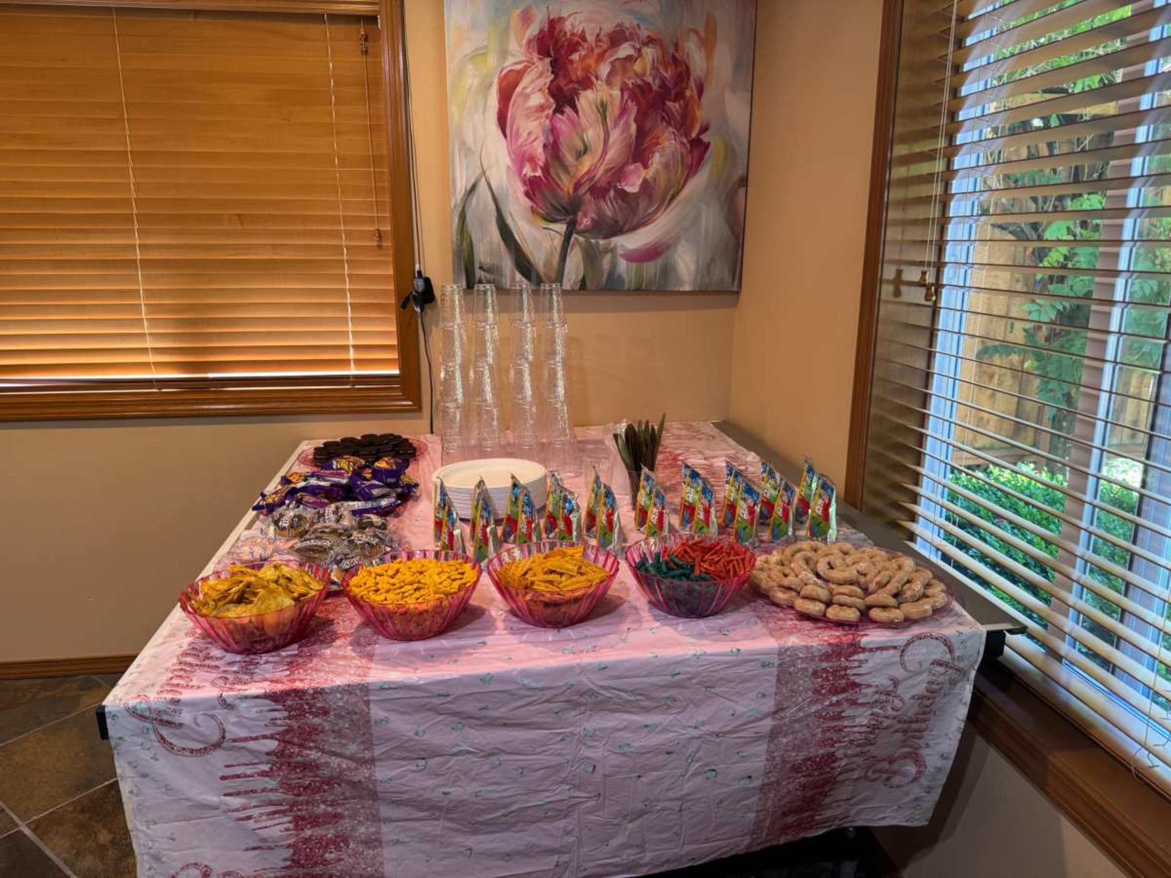 A table covered with a pink cloth displays an assortment of snacks, including bowls of chips, candy, pretzels, and cookies, alongside plastic cups and decorative items.