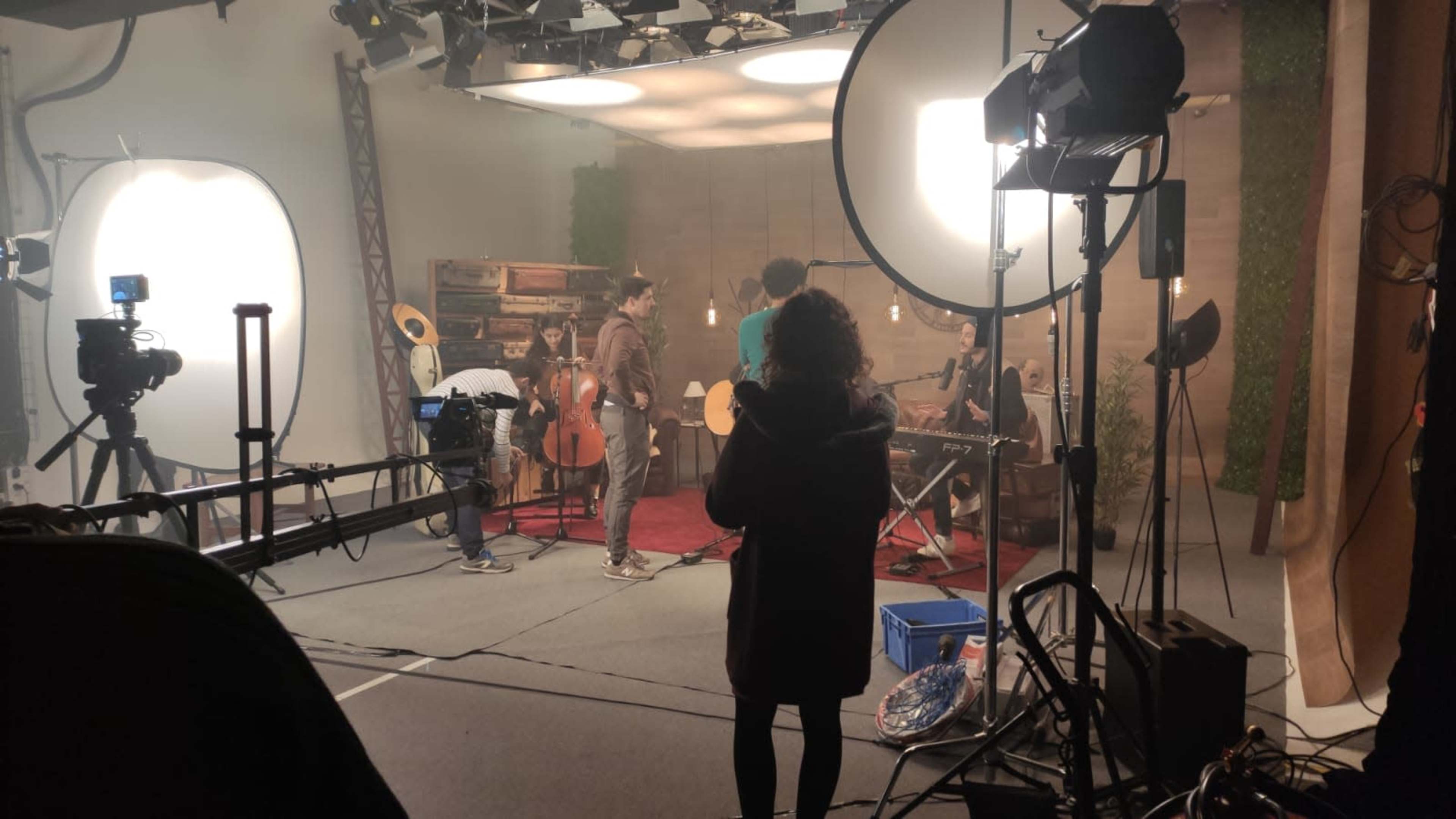 A group of musicians is rehearsing in a studio, surrounded by cameras and lighting equipment.