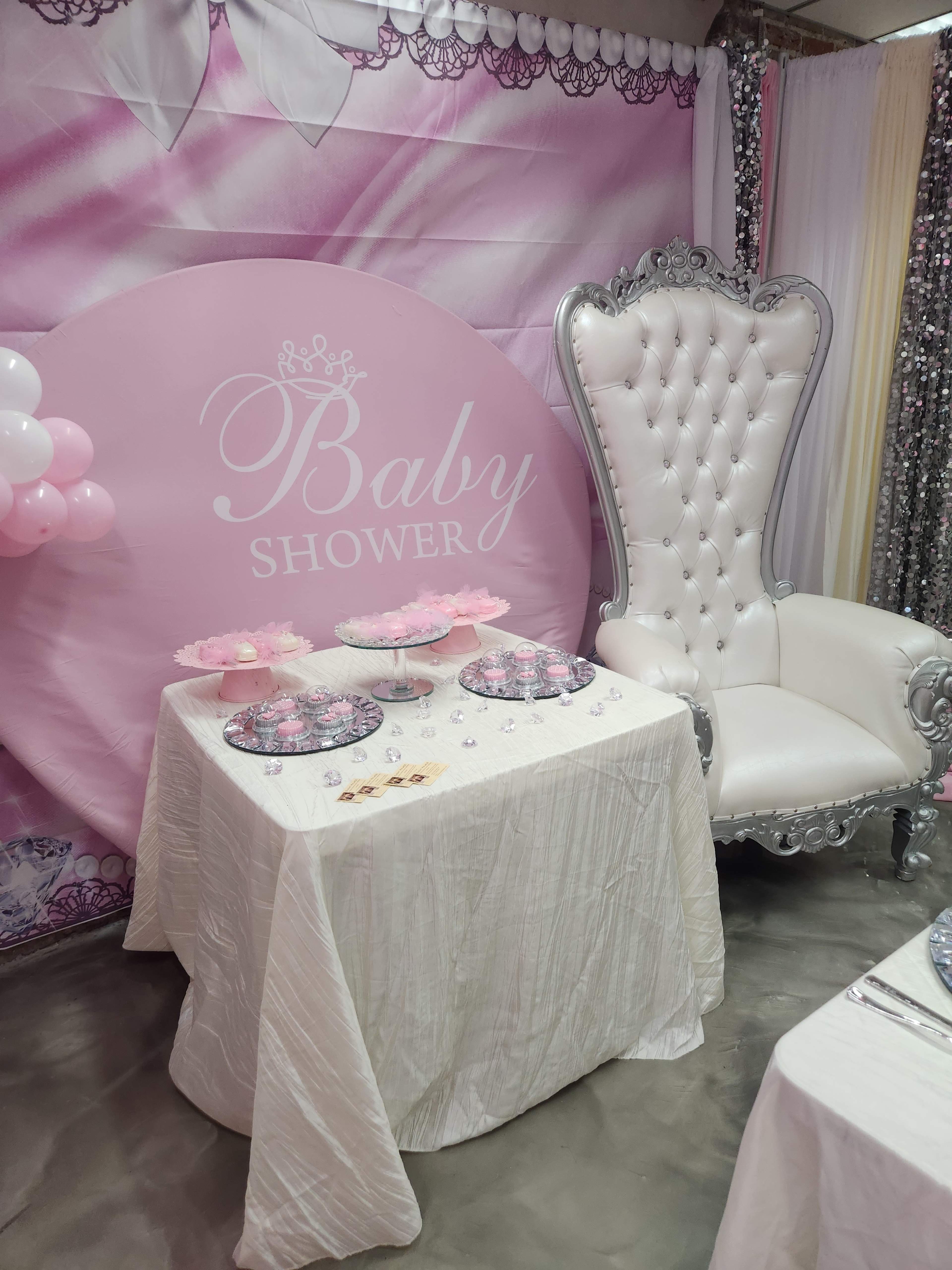 A decorated baby shower setting with a large pink backdrop, a white table covered with a cloth, and an ornate silver throne chair.