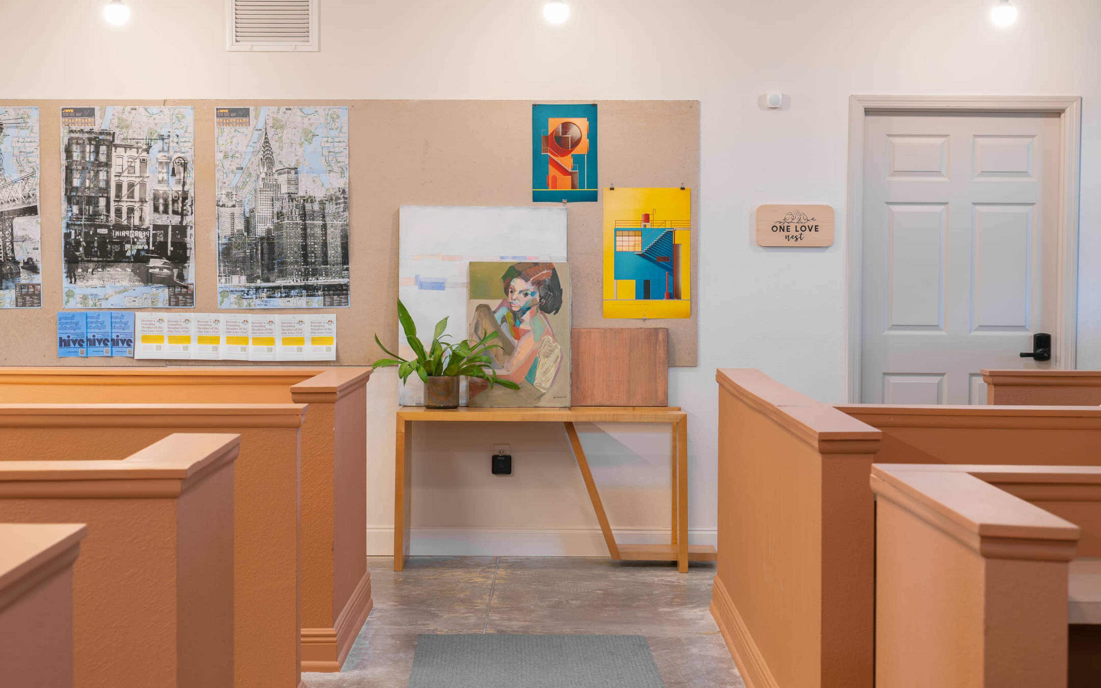 The image shows a well-lit interior of a space with wooden benches, a table displaying artwork and a plant, and walls featuring framed art and a bulletin board.
