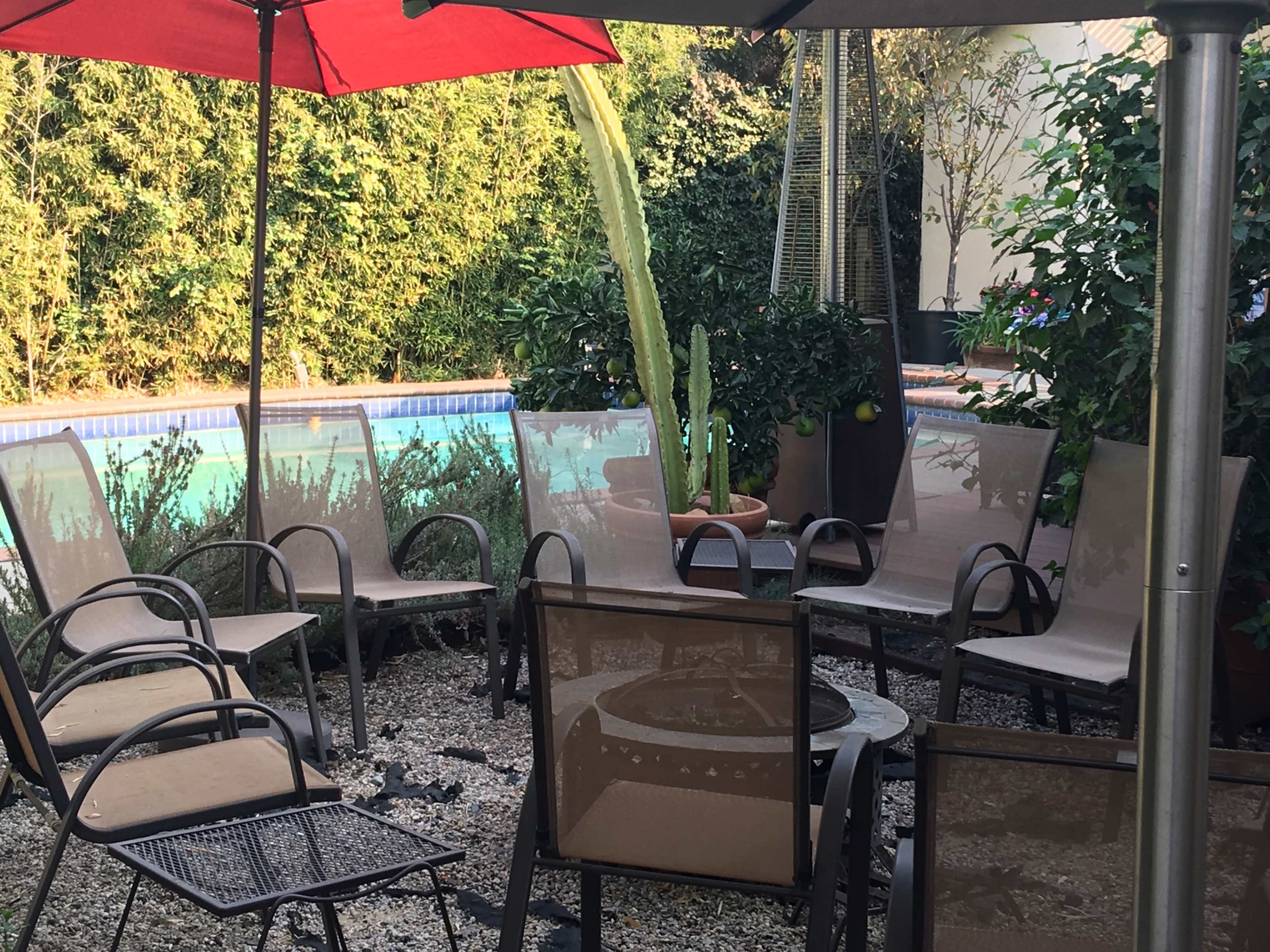 A patio area with several metal and fabric chairs arranged around a table, surrounded by greenery and a swimming pool in the background.
