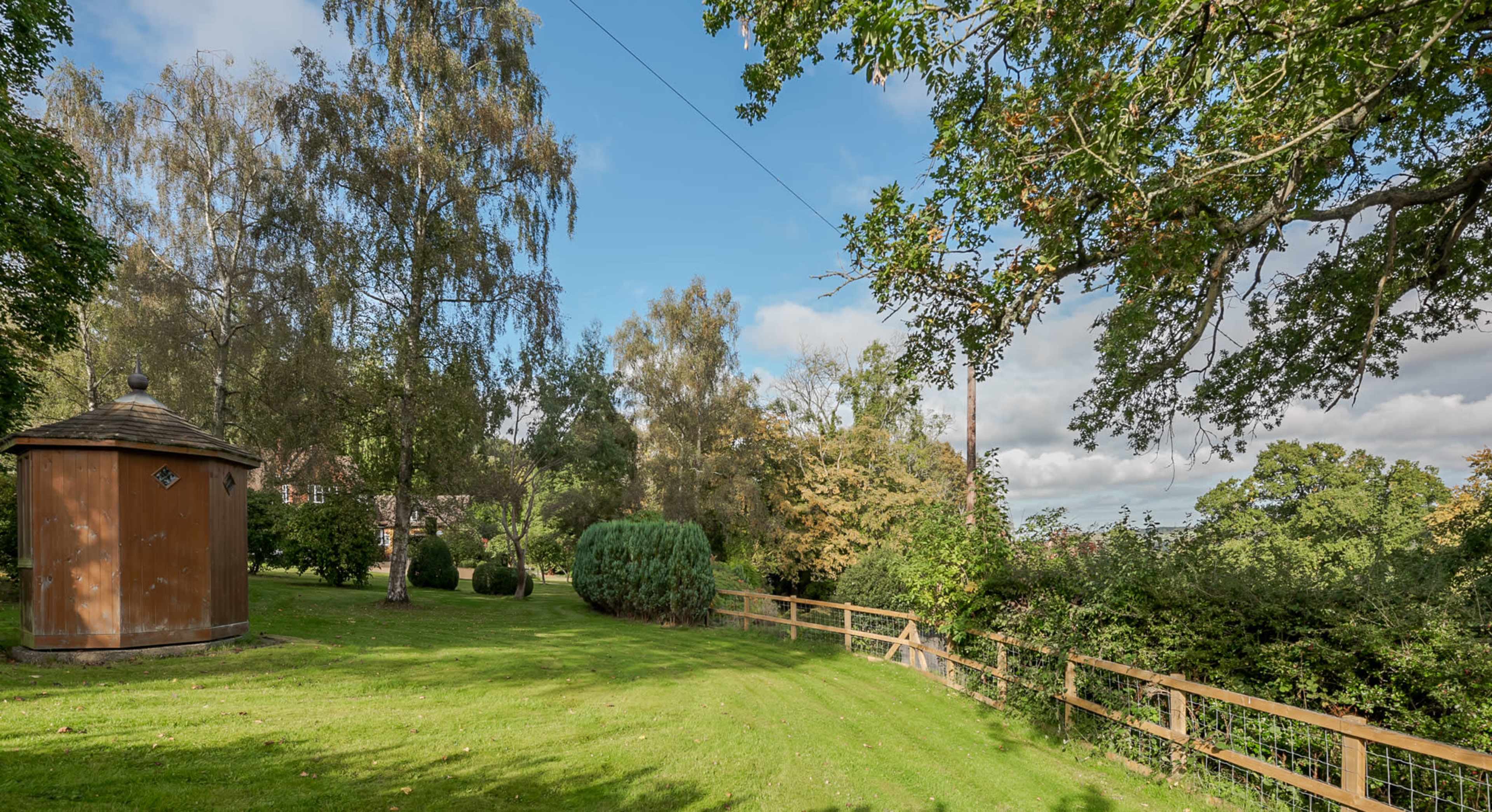 A green lawn bordered by bushes and trees, with a wooden structure and a fence in the background under a partly cloudy sky.