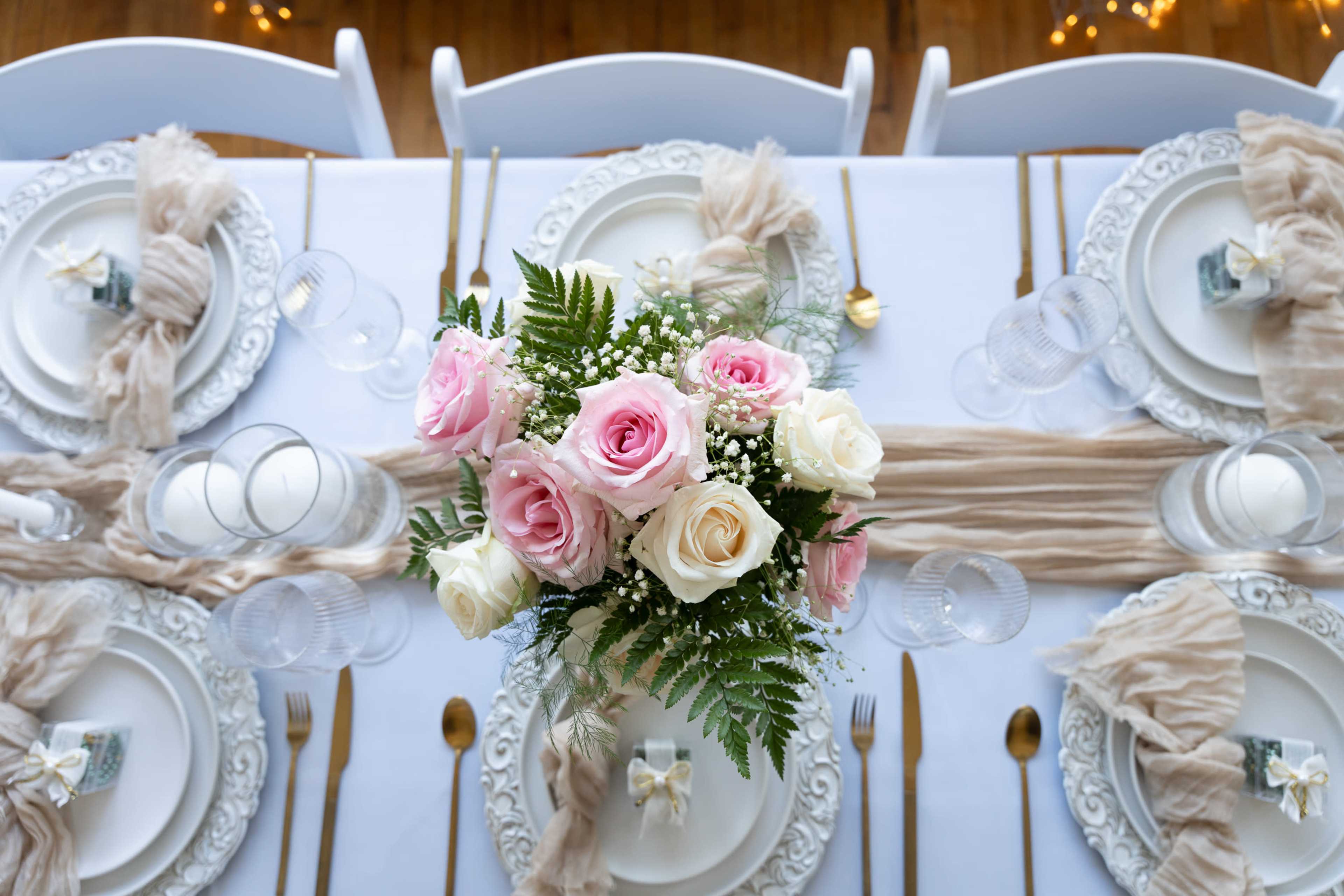 A dining table is set with white plates, gold utensils, clear glasses, and a centerpiece of pink and white roses surrounded by greenery.