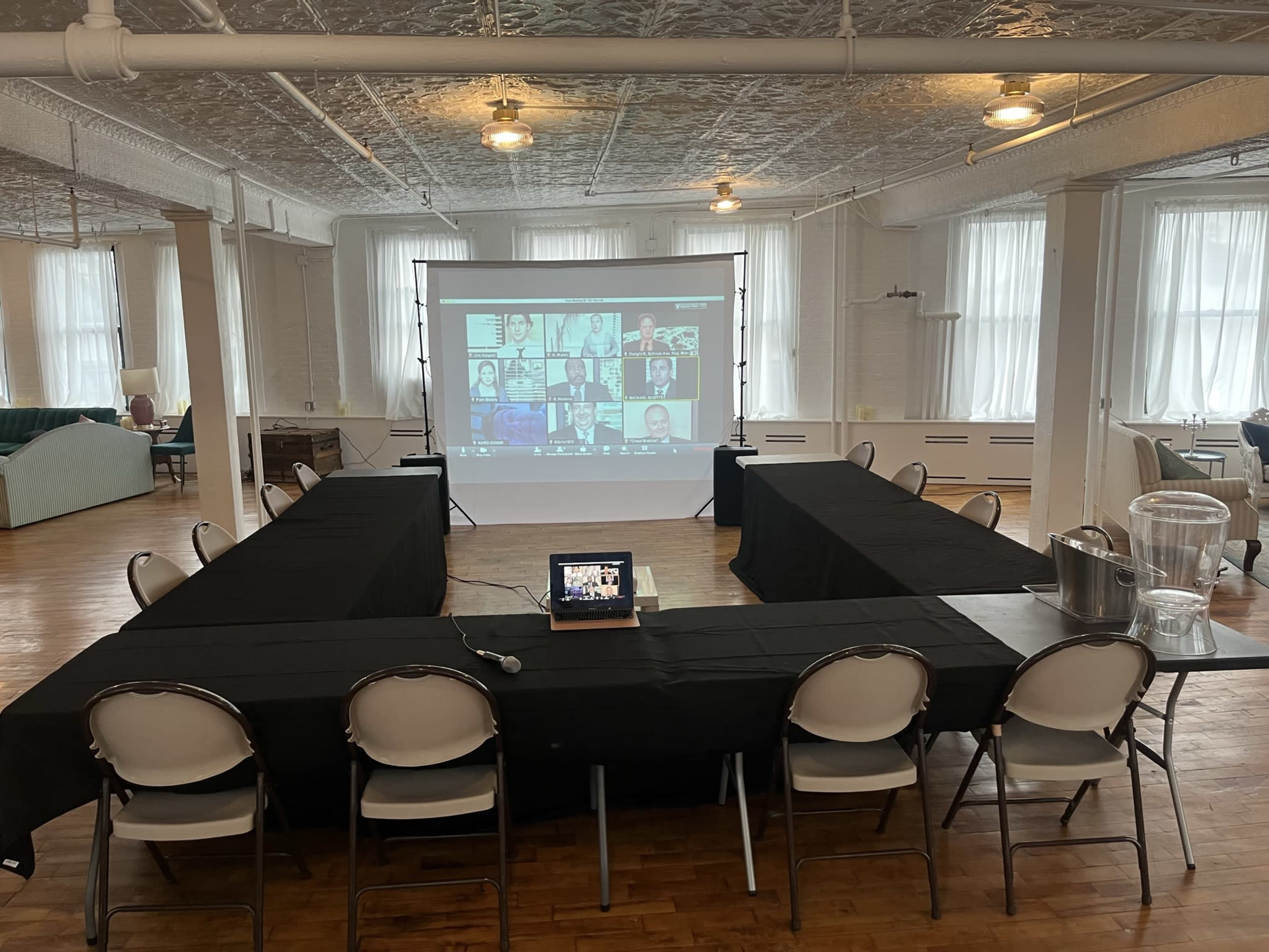 A large conference room is set up with a rectangular arrangement of tables covered in black cloth, facing a screen displaying a video call.