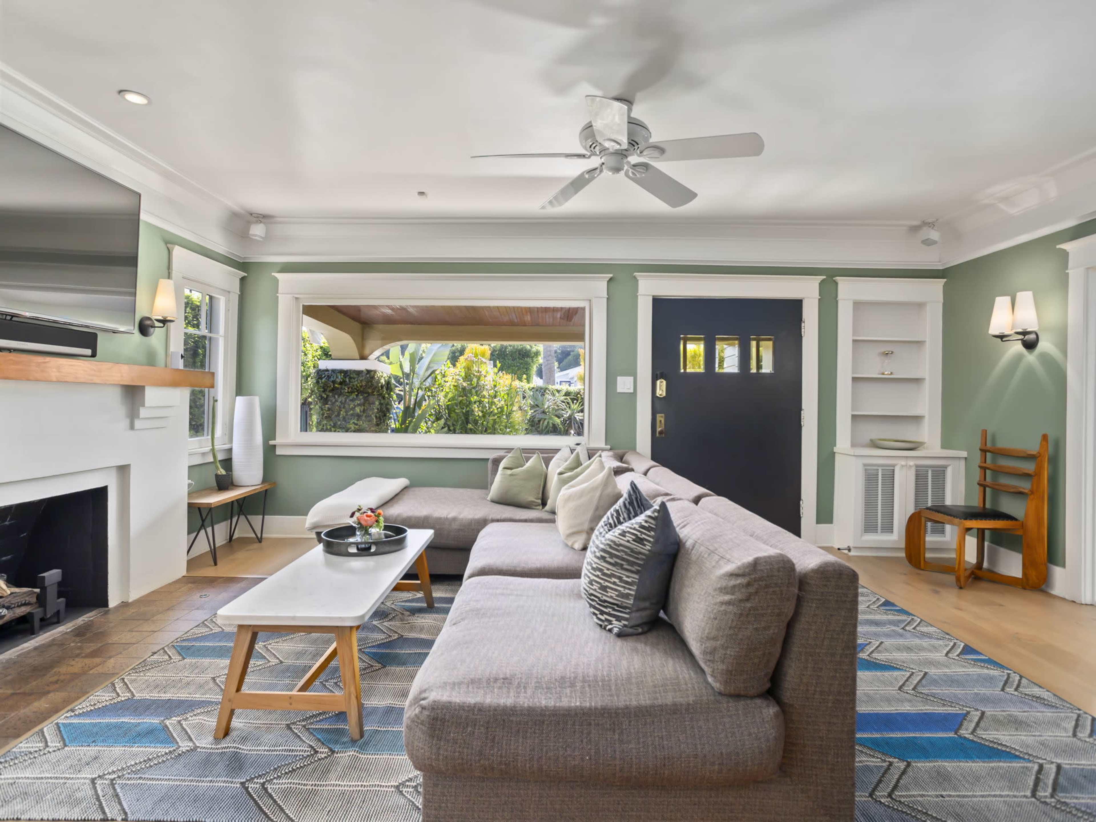 The image shows a well-lit living room featuring a gray sectional sofa, a coffee table, a modern ceiling fan, and a view of a garden through a large window.