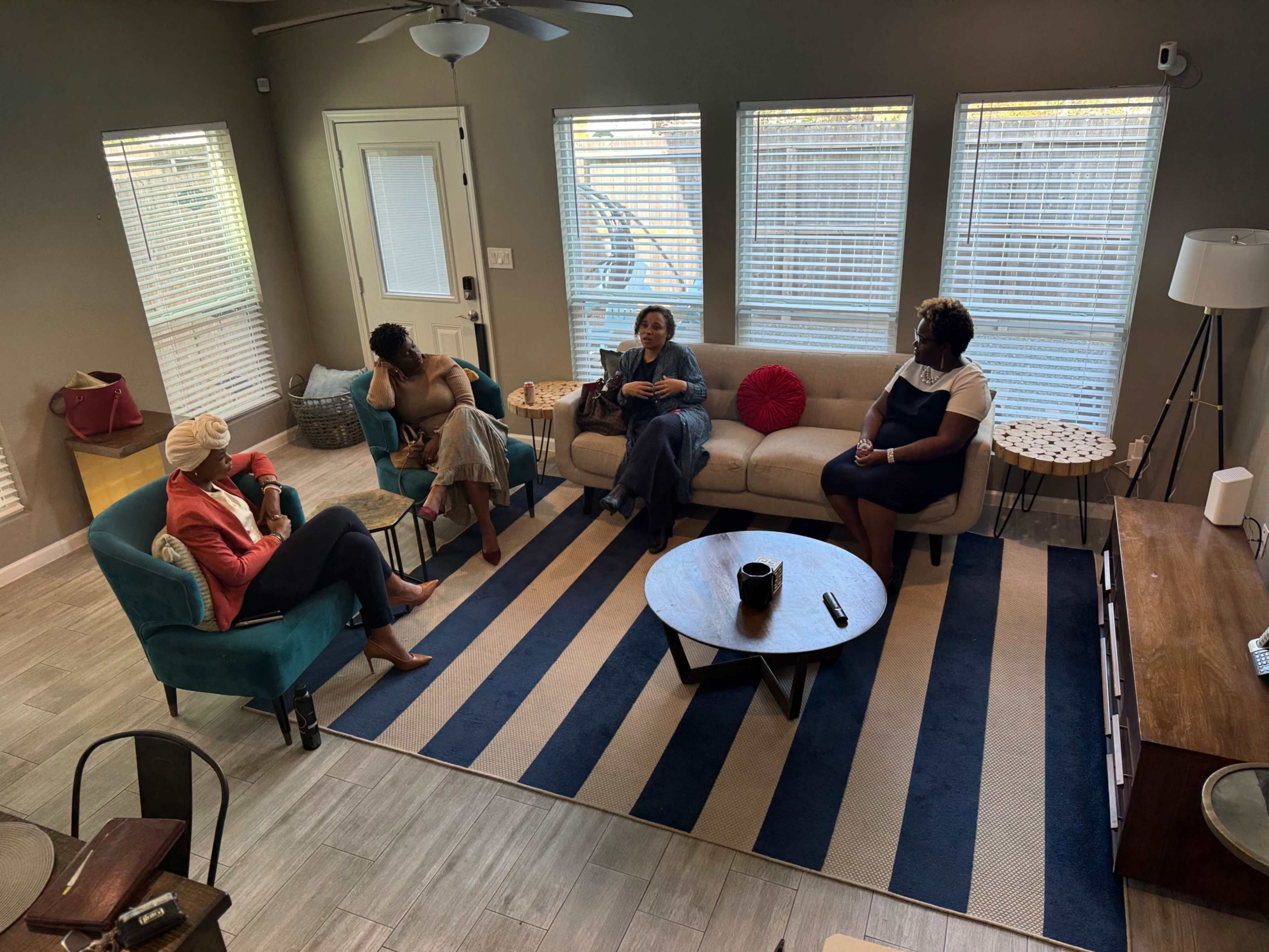 Four women sit in a living room, arranged on a sofa and chairs around a coffee table on a striped rug.