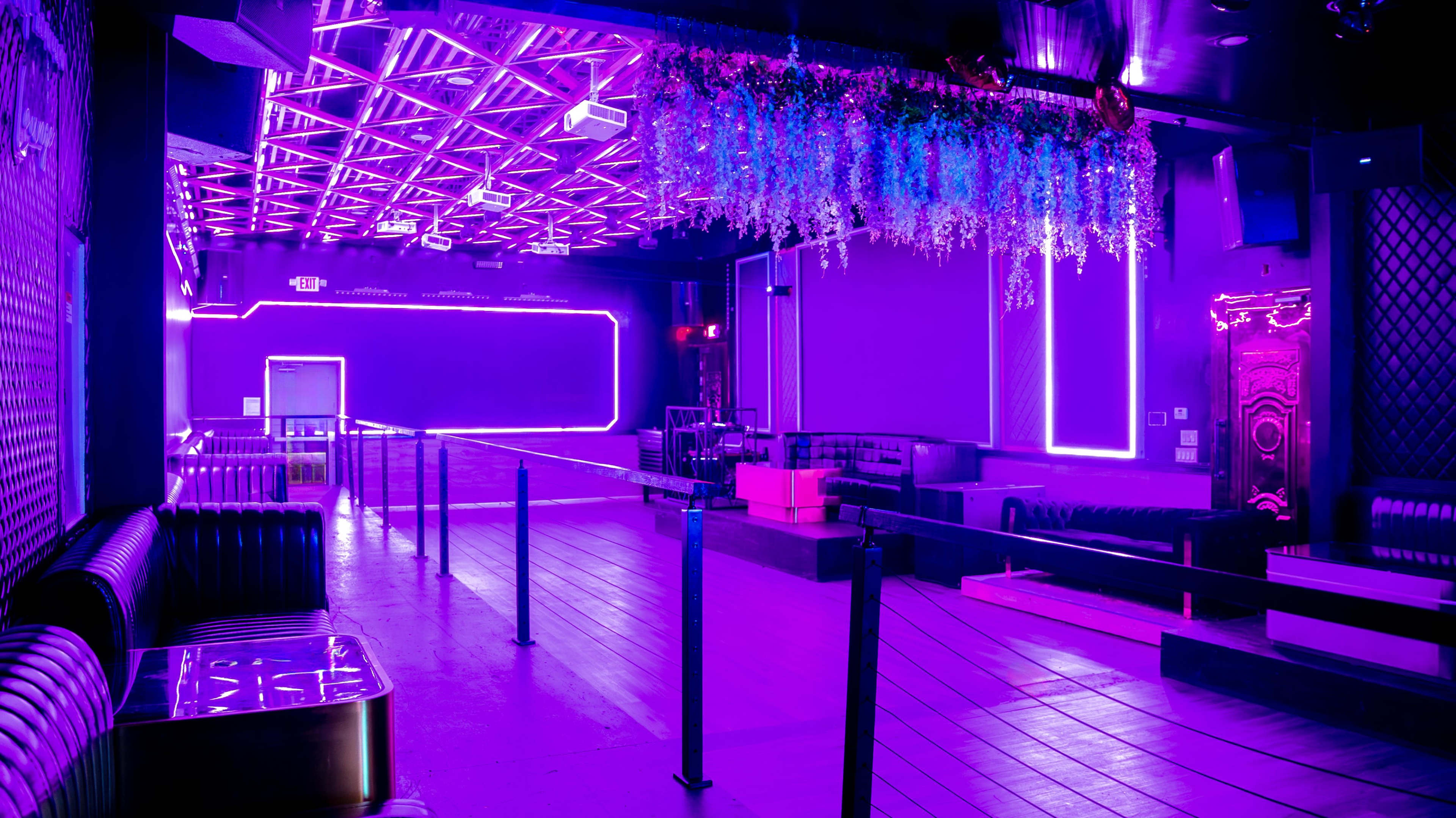 The image shows a dimly lit nightclub interior with purple neon lighting and a row of empty seating areas.