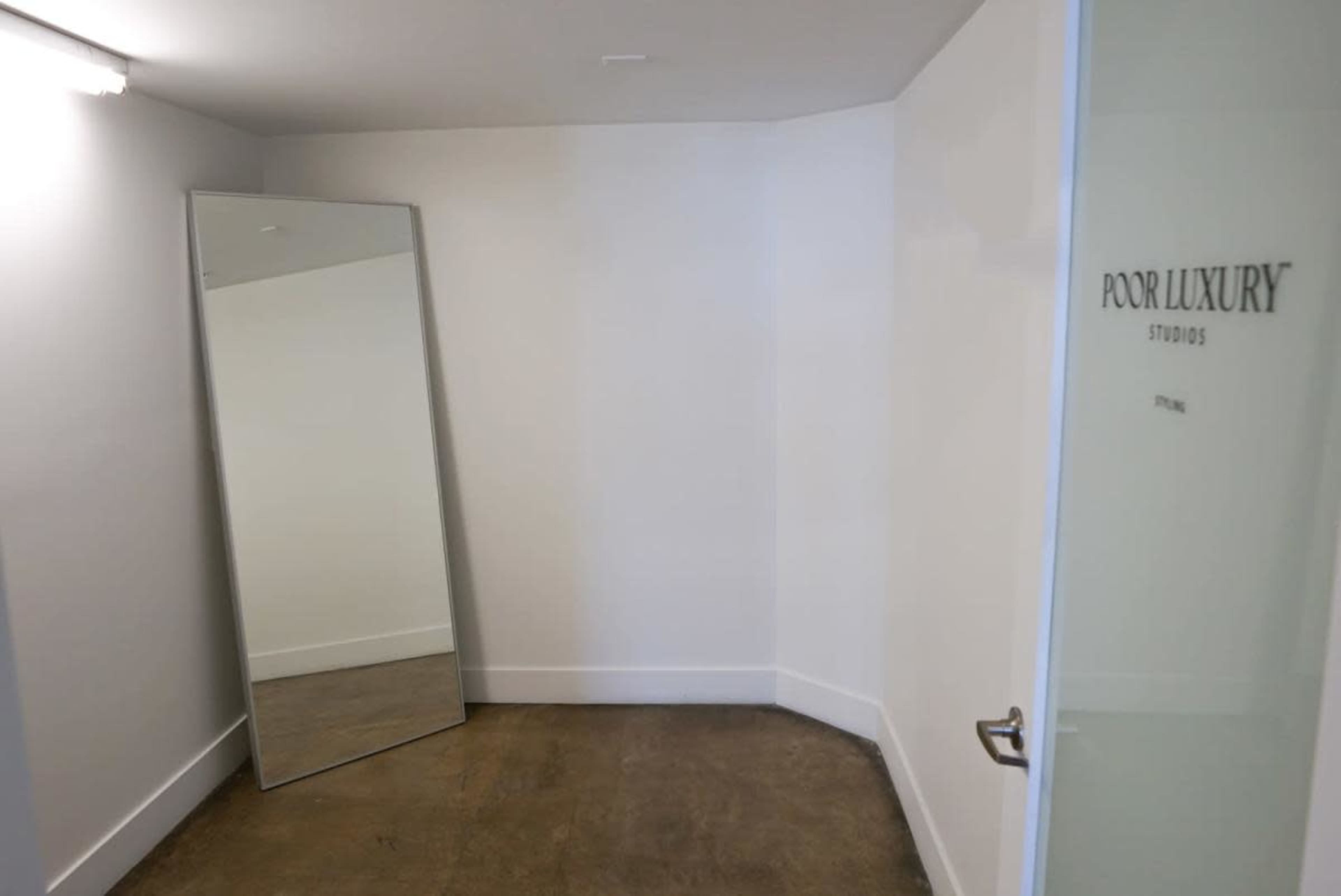 The image shows a minimalist room with a large mirror leaning against the wall and a glass door labeled "POOR LUXURY STUDIOS."