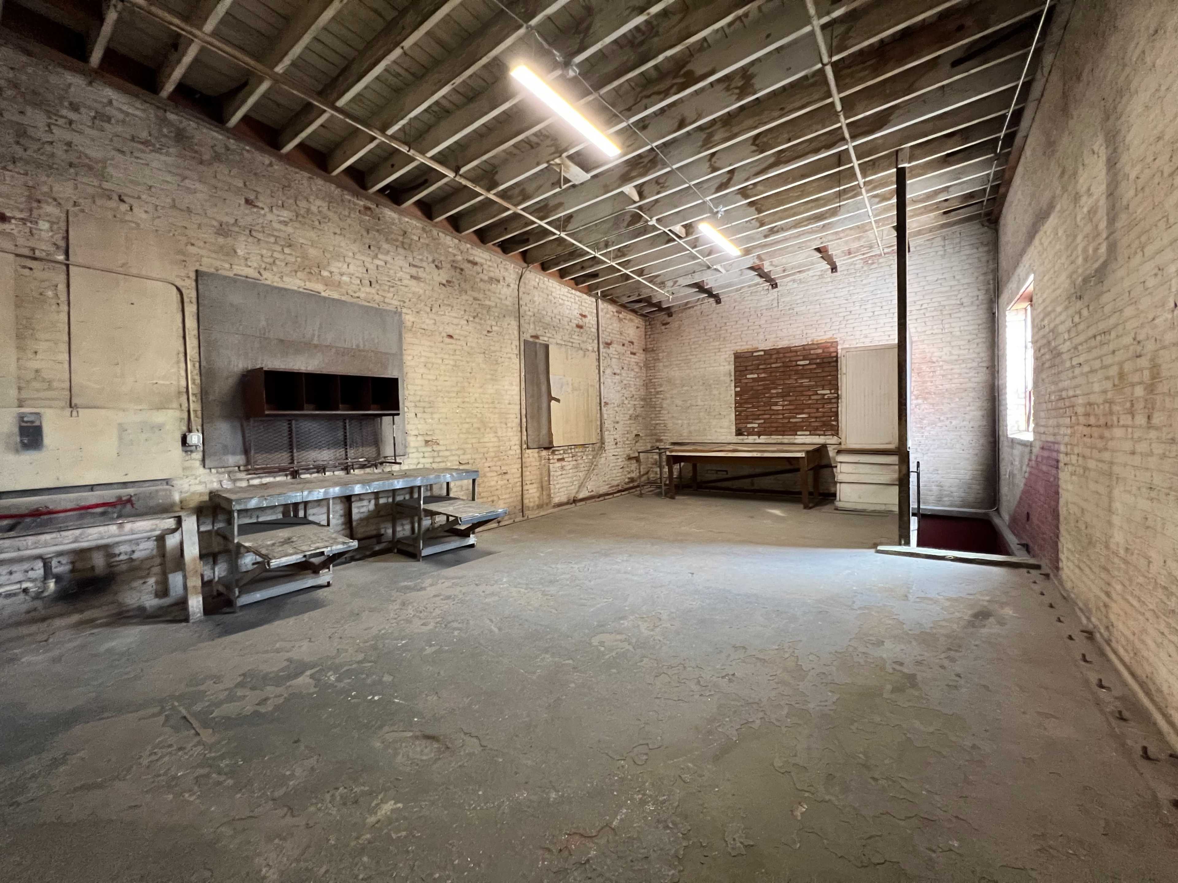 An empty, industrial-style room with exposed brick walls, wooden beams, and minimal furniture, including a couple of worktables and a staircase.