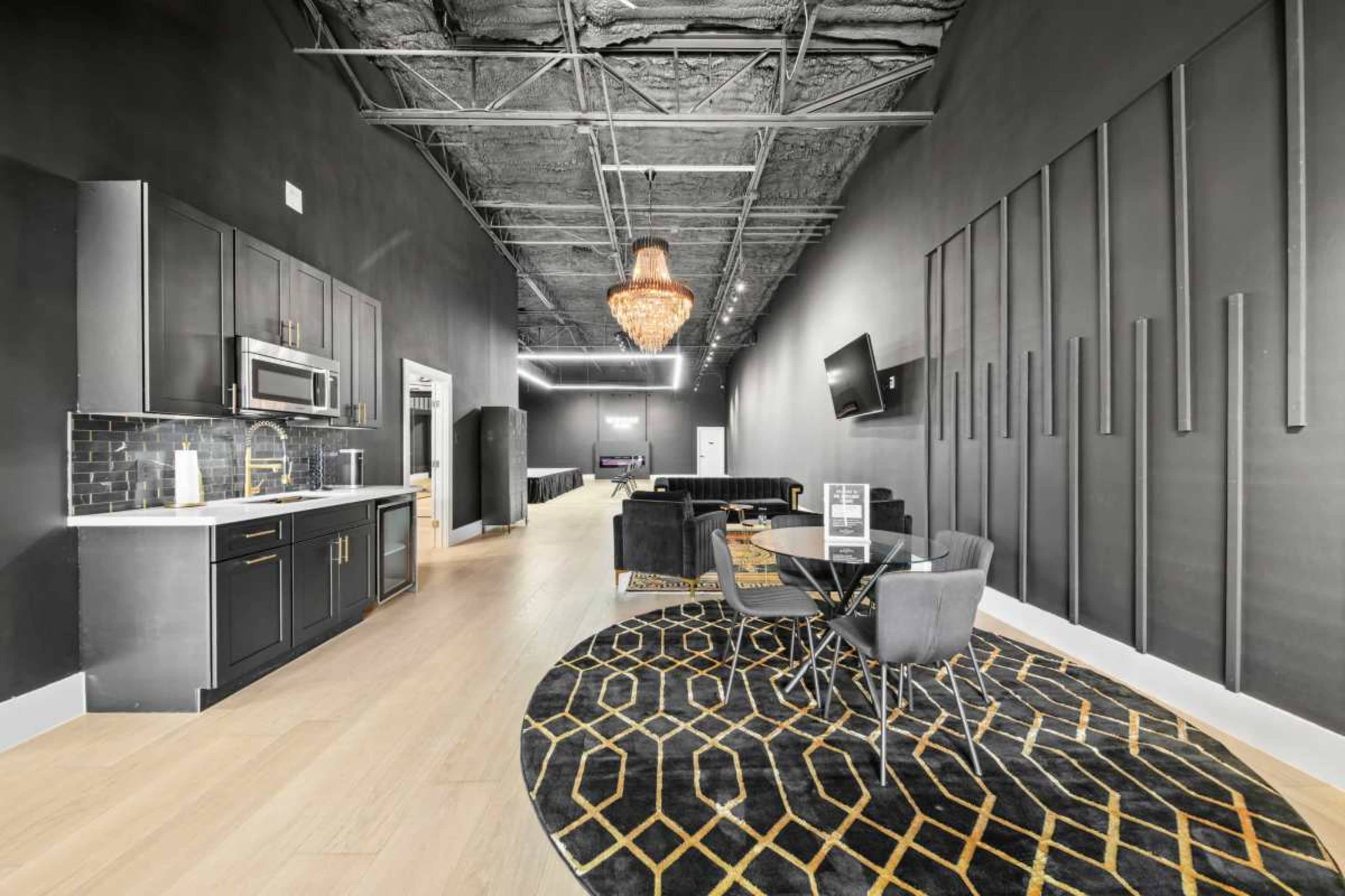 The image shows a modern interior space featuring a kitchen area with dark cabinetry, a dining table with chairs, and a chandelier overhead, all set against a black and gold color scheme.