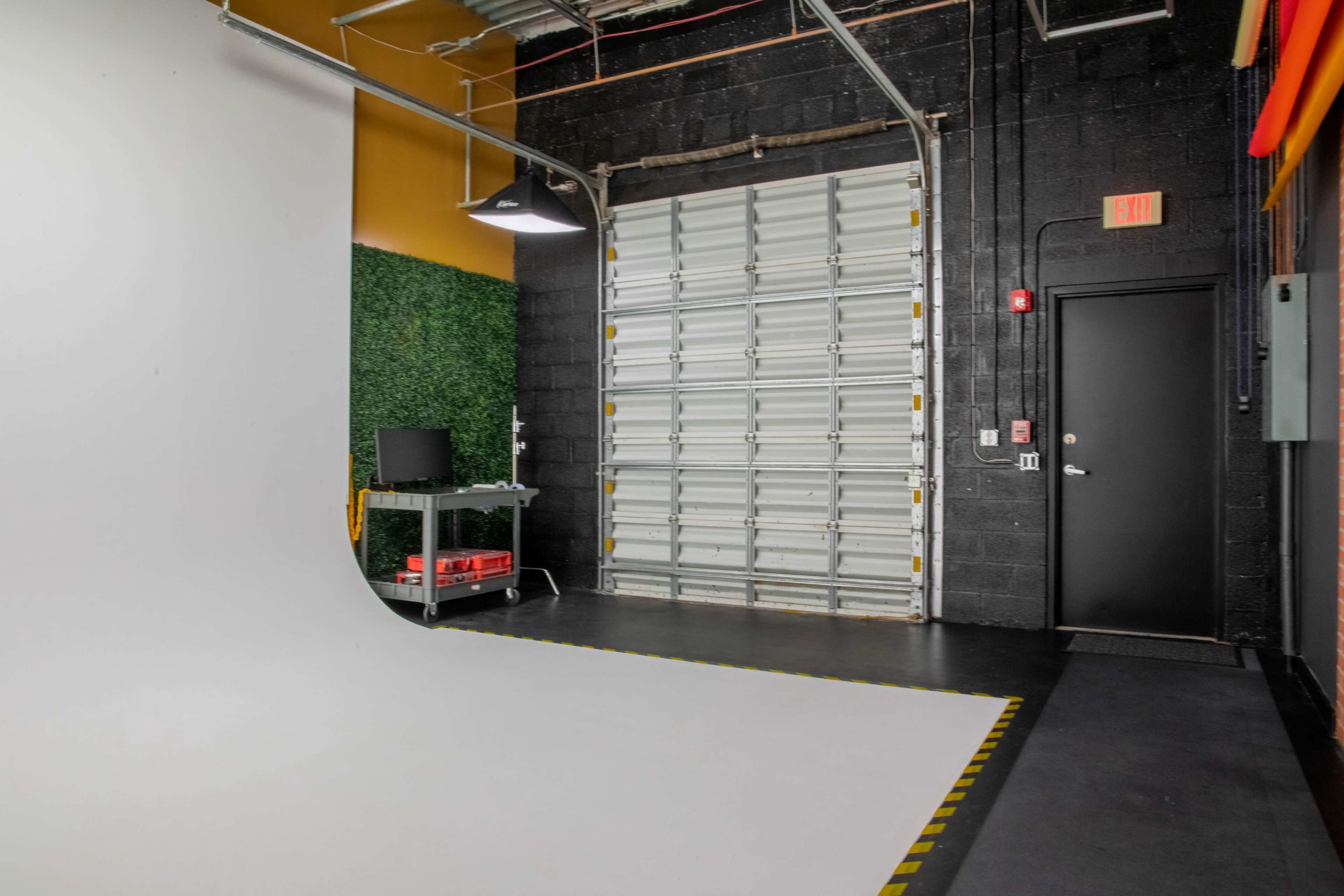 A photography studio with a seamless white backdrop, a green wall, a rolling door, and a small workstation with a monitor in the corner.