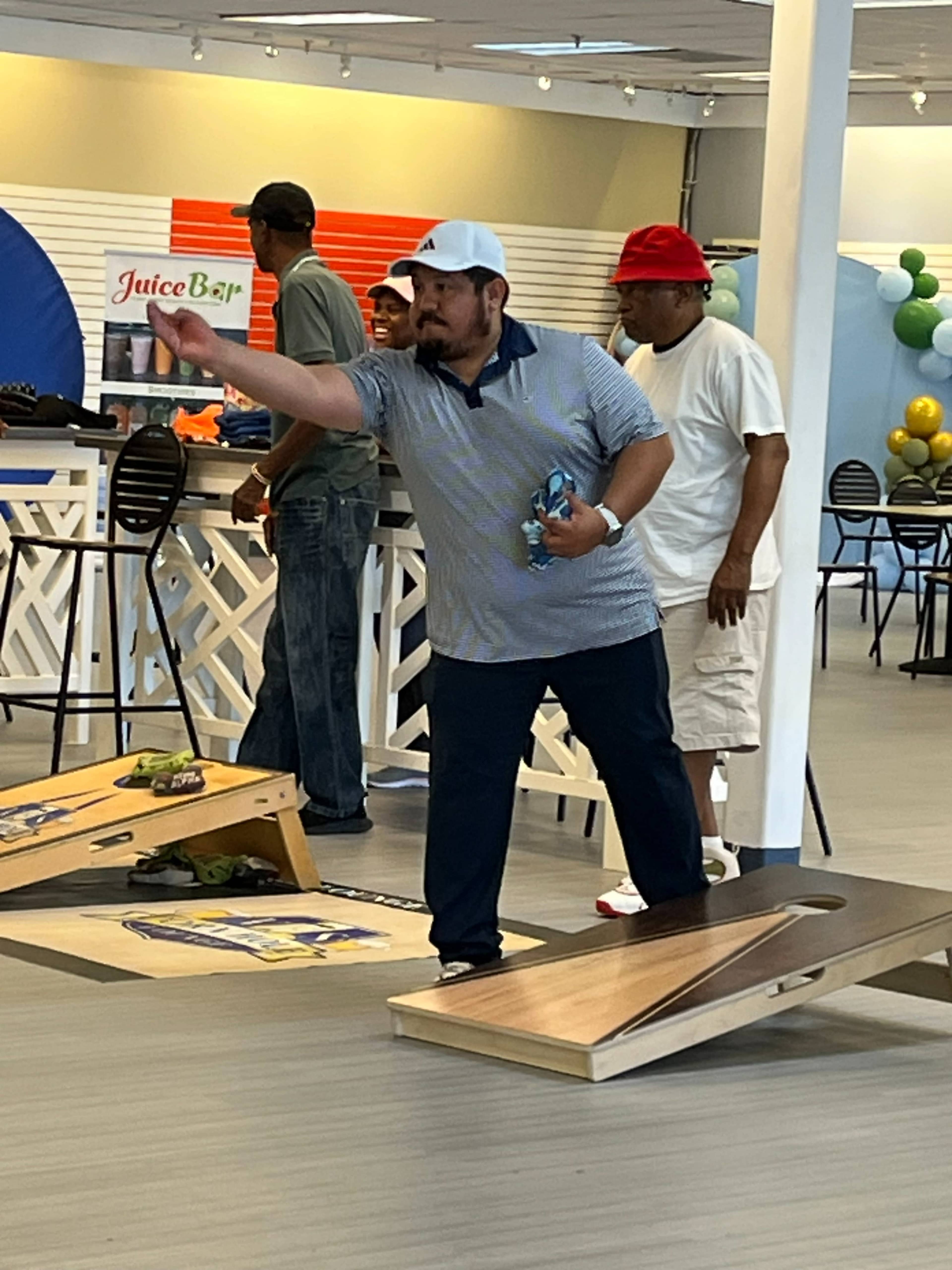 A man wearing a golf cap throws a bean bag towards a cornhole board in a recreational space, while others stand nearby.