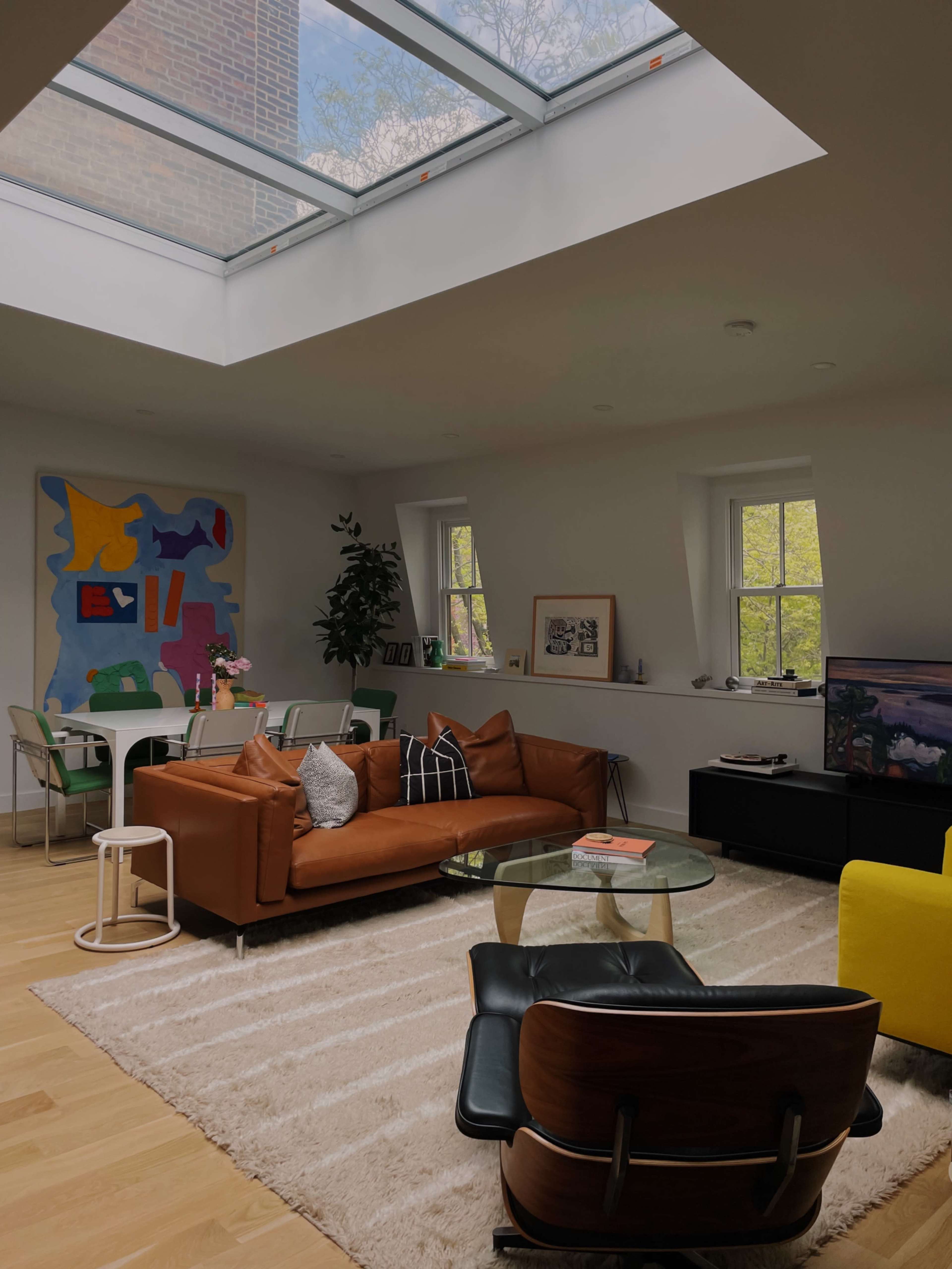The image shows a modern living room with a large skylight, a brown leather sofa, and a round glass coffee table, surrounded by colorful art and furnishings.