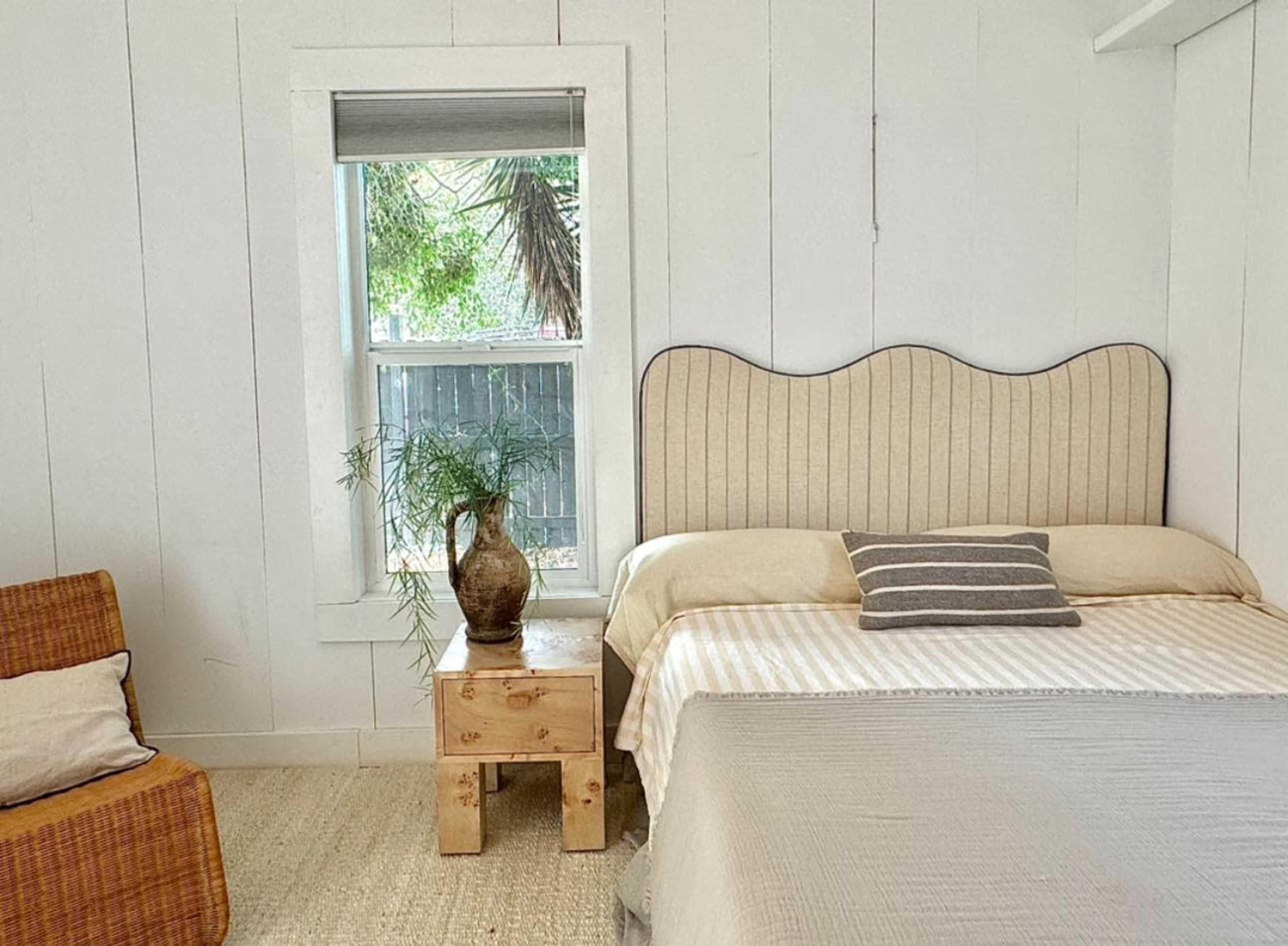 A bed with a headboard and a small wooden table sits next to a window in a light-colored room.