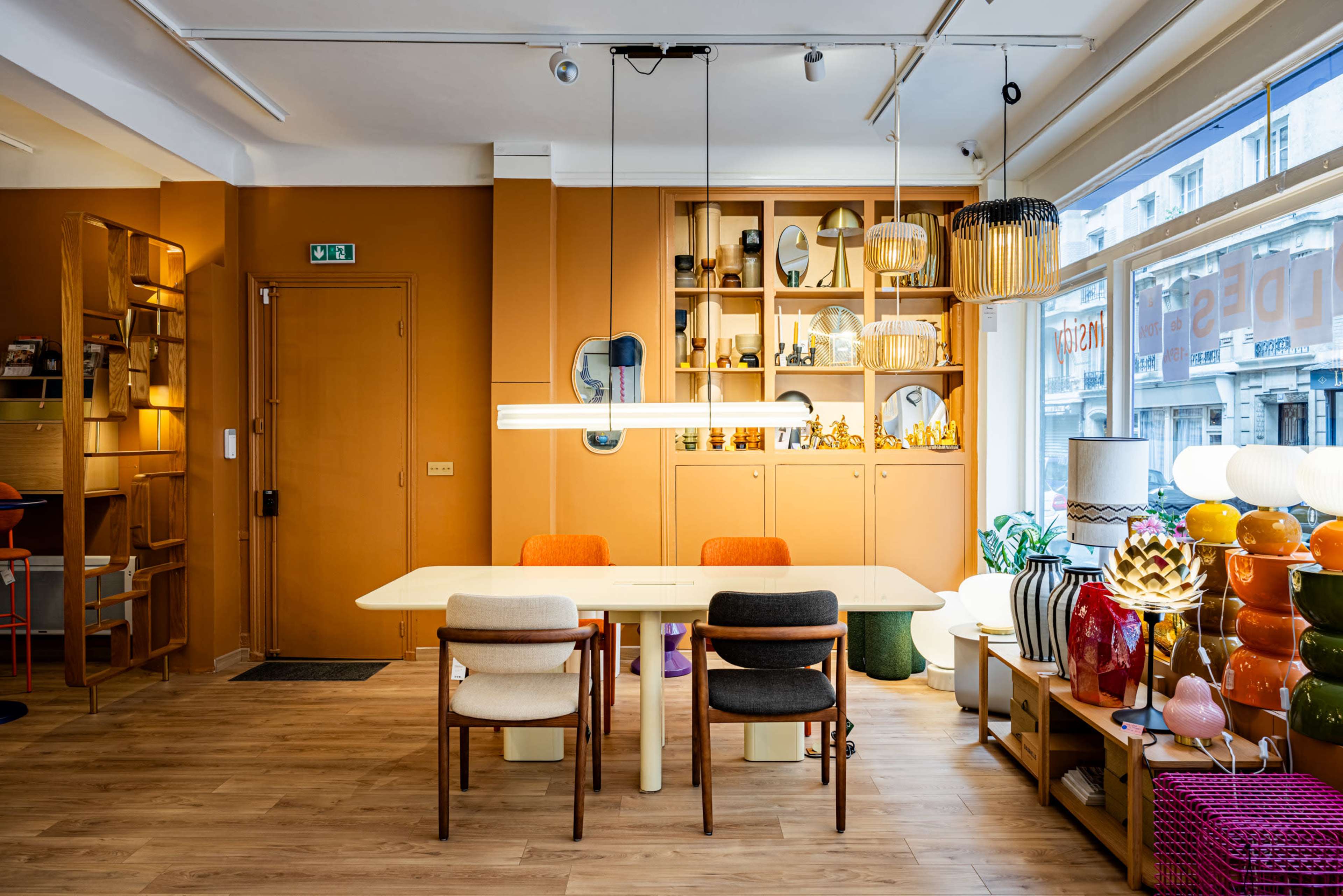 The image shows a modern interior design of a room featuring a dining table with four chairs, a well-organized shelf displaying decorative items, and various light fixtures.